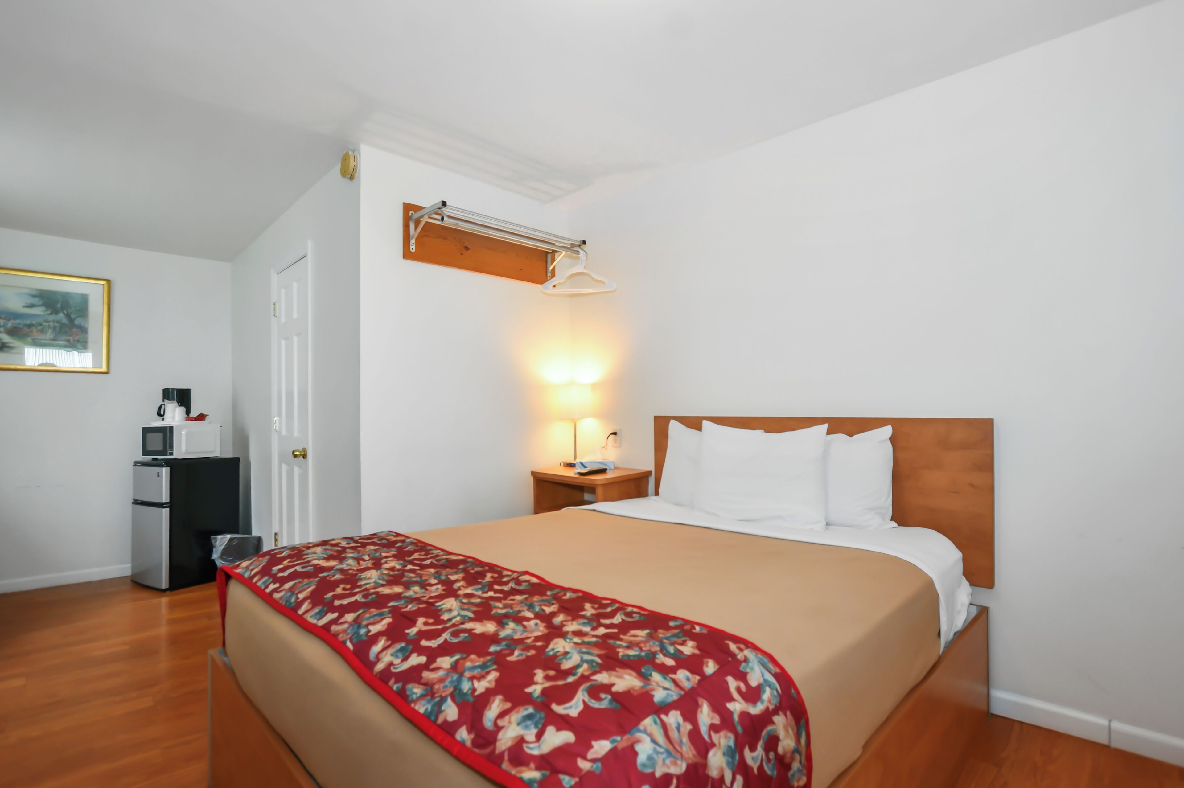 A hotel room with a bed, a nightstand with a lamp, a coat rack, a microwave, and a refrigerator.