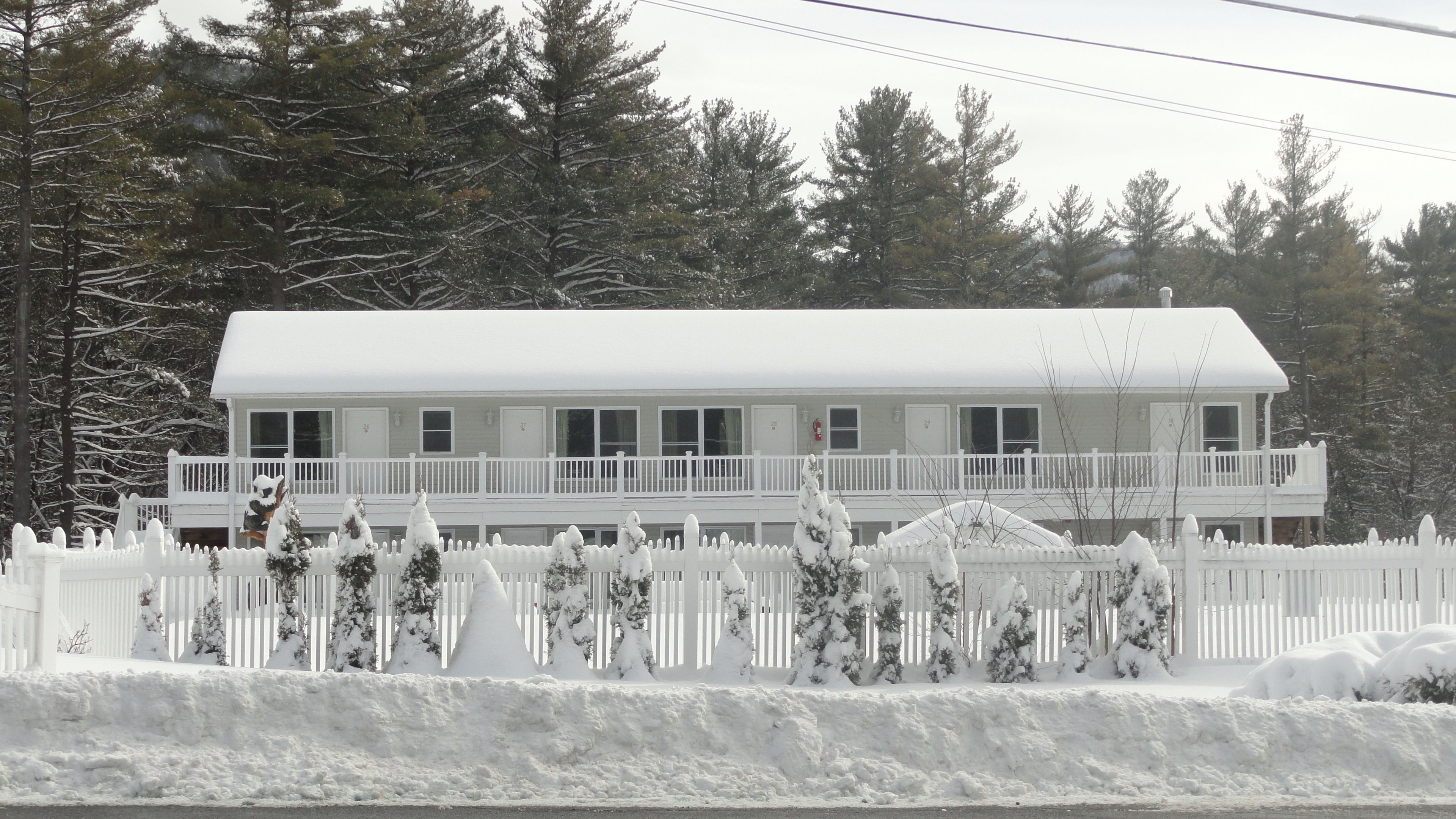 A two-story white building with a balcony, surrounded by a snow-covered fence and trees.