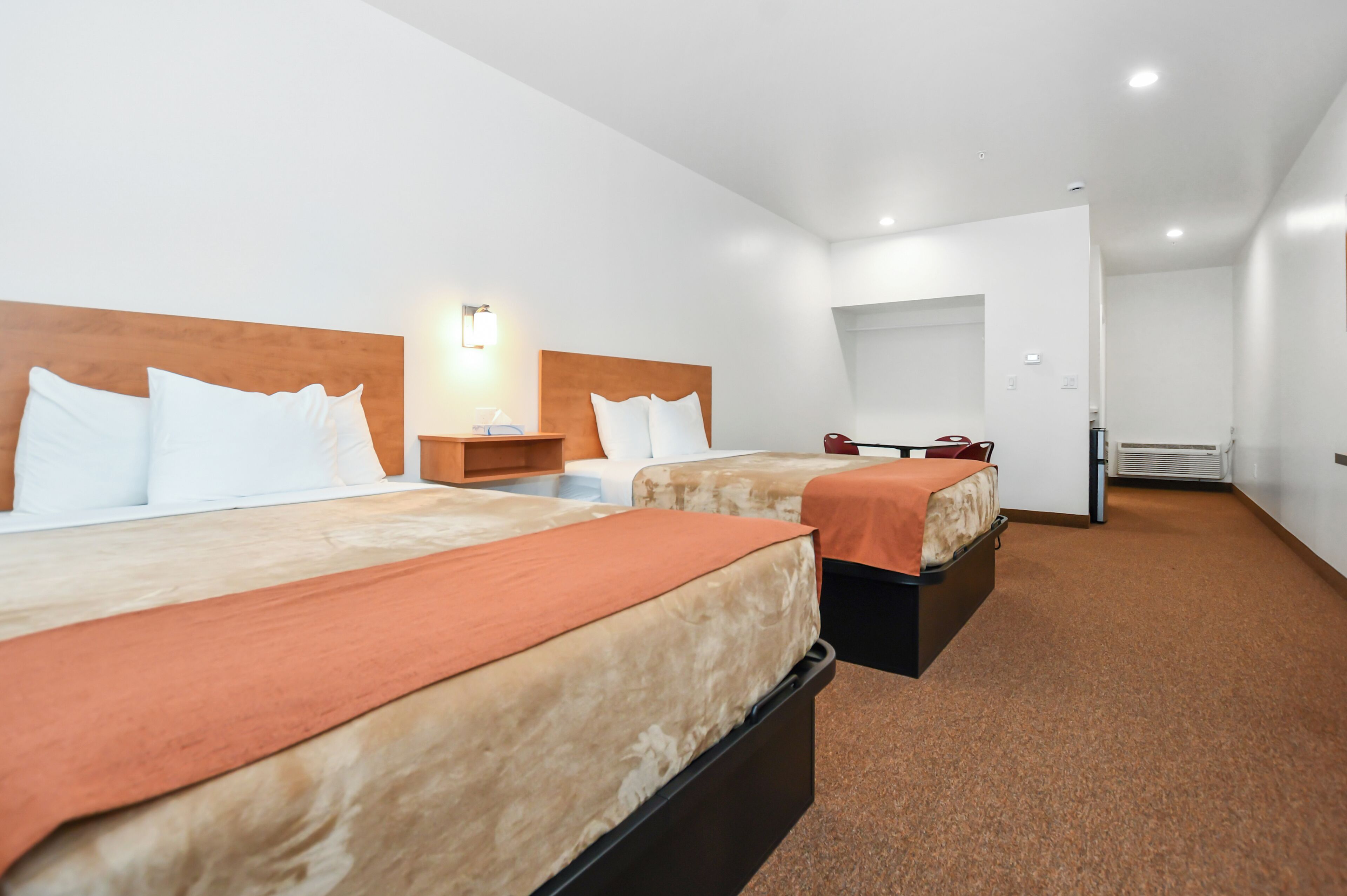 A hotel room with two beds, a wooden headboard, a nightstand, a lamp, and a door leading to a bathroom.