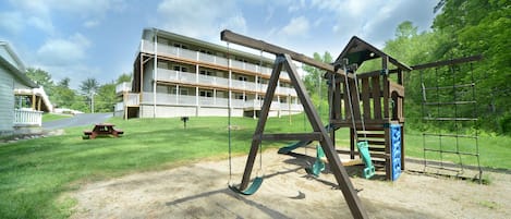 A playground with swings and a picnic table in front of a multi-story building.
