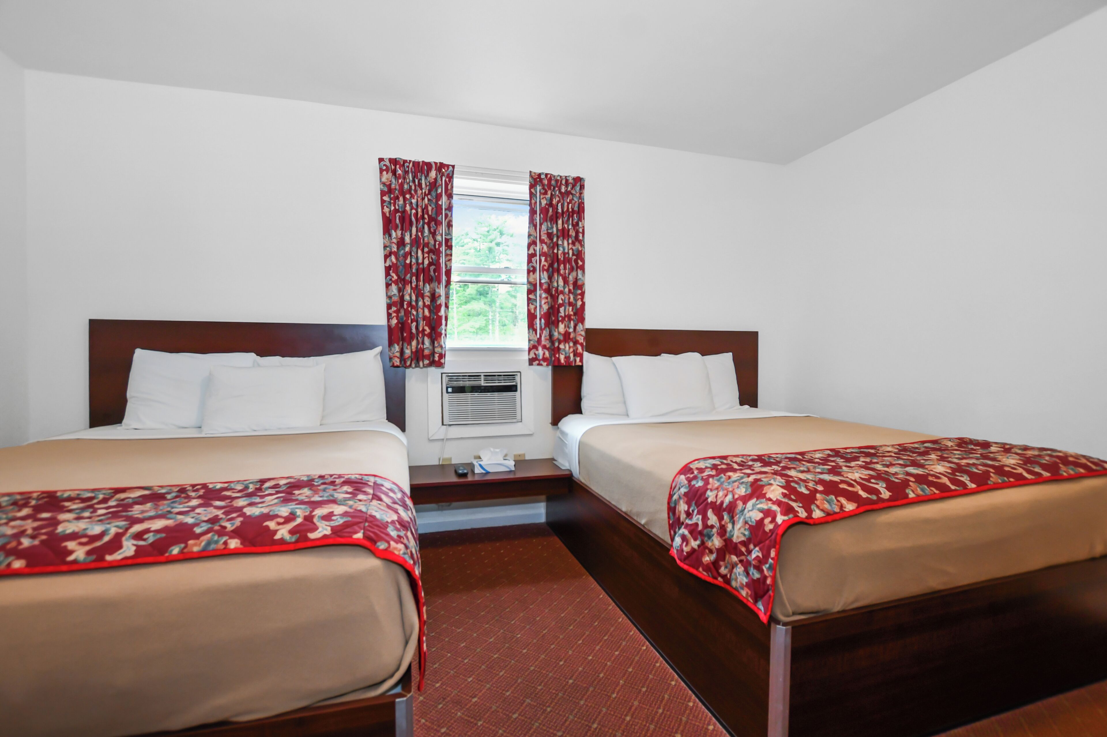 A hotel room with two beds, a window with curtains, an air conditioning unit, and a small table.