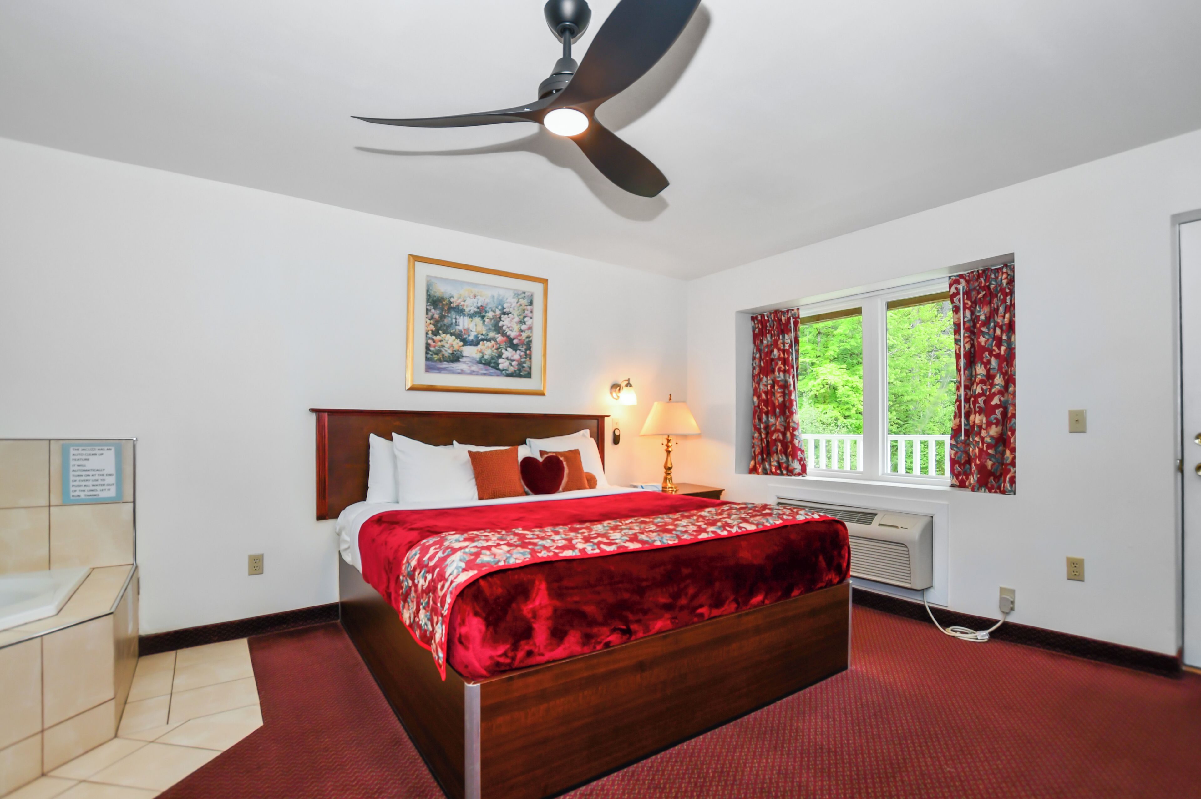Habitación de hotel con cama, ventilador de techo, ventana con cortinas, un cuadro en la pared y una lámpara de noche.