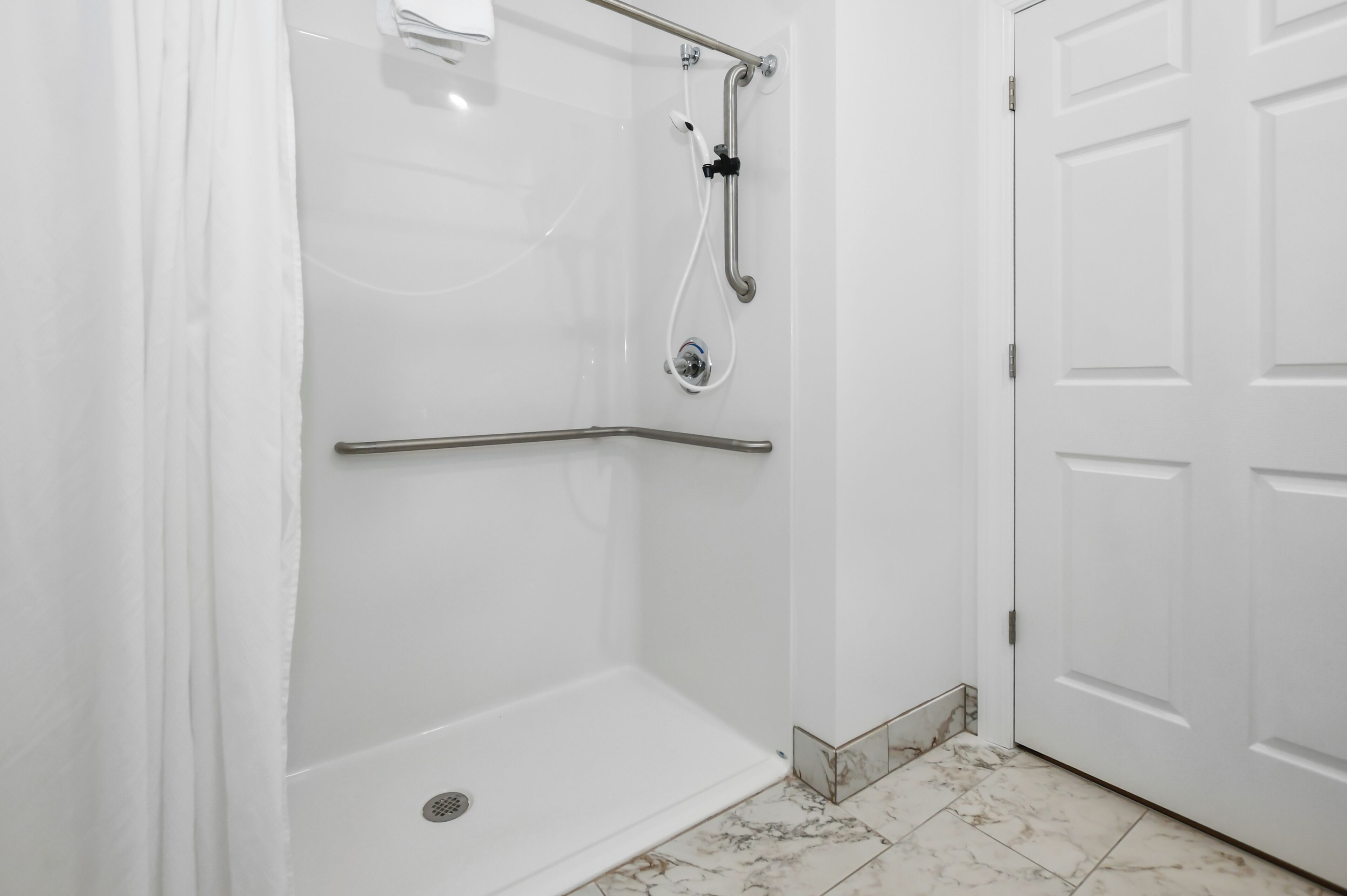 A white shower enclosure with a grab bar, a door, and a marble-tiled floor.