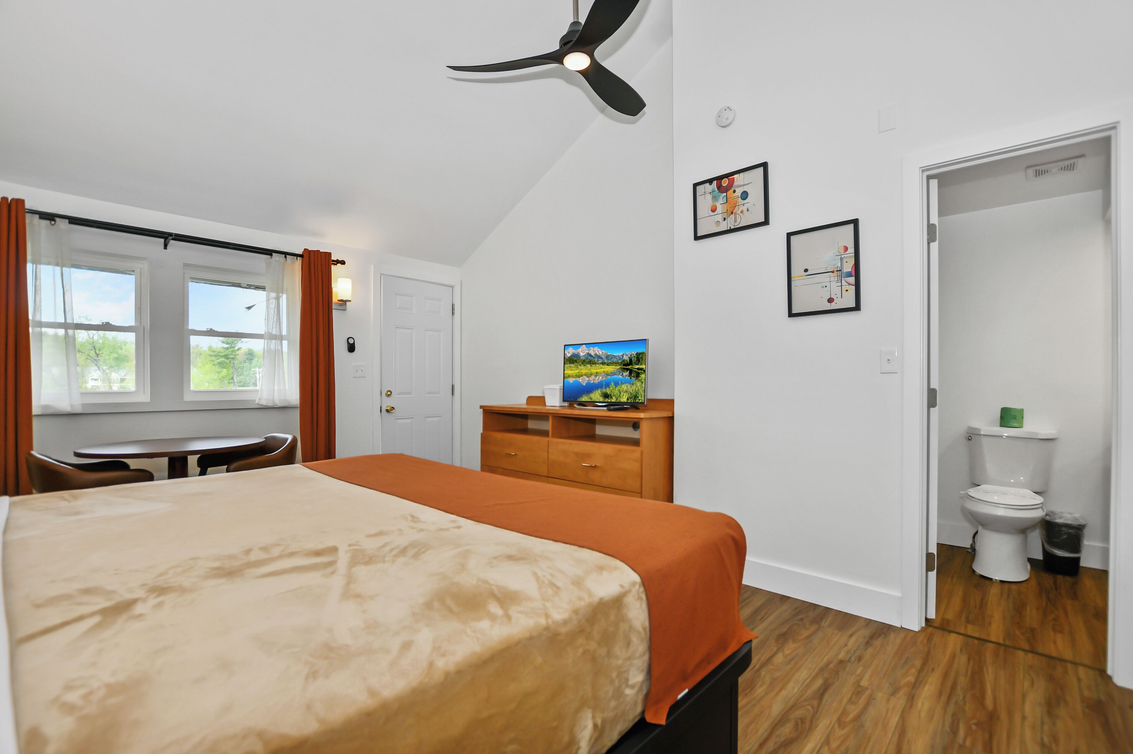 A bedroom with a bed, a desk, a chair, a ceiling fan, a TV, and a toilet.