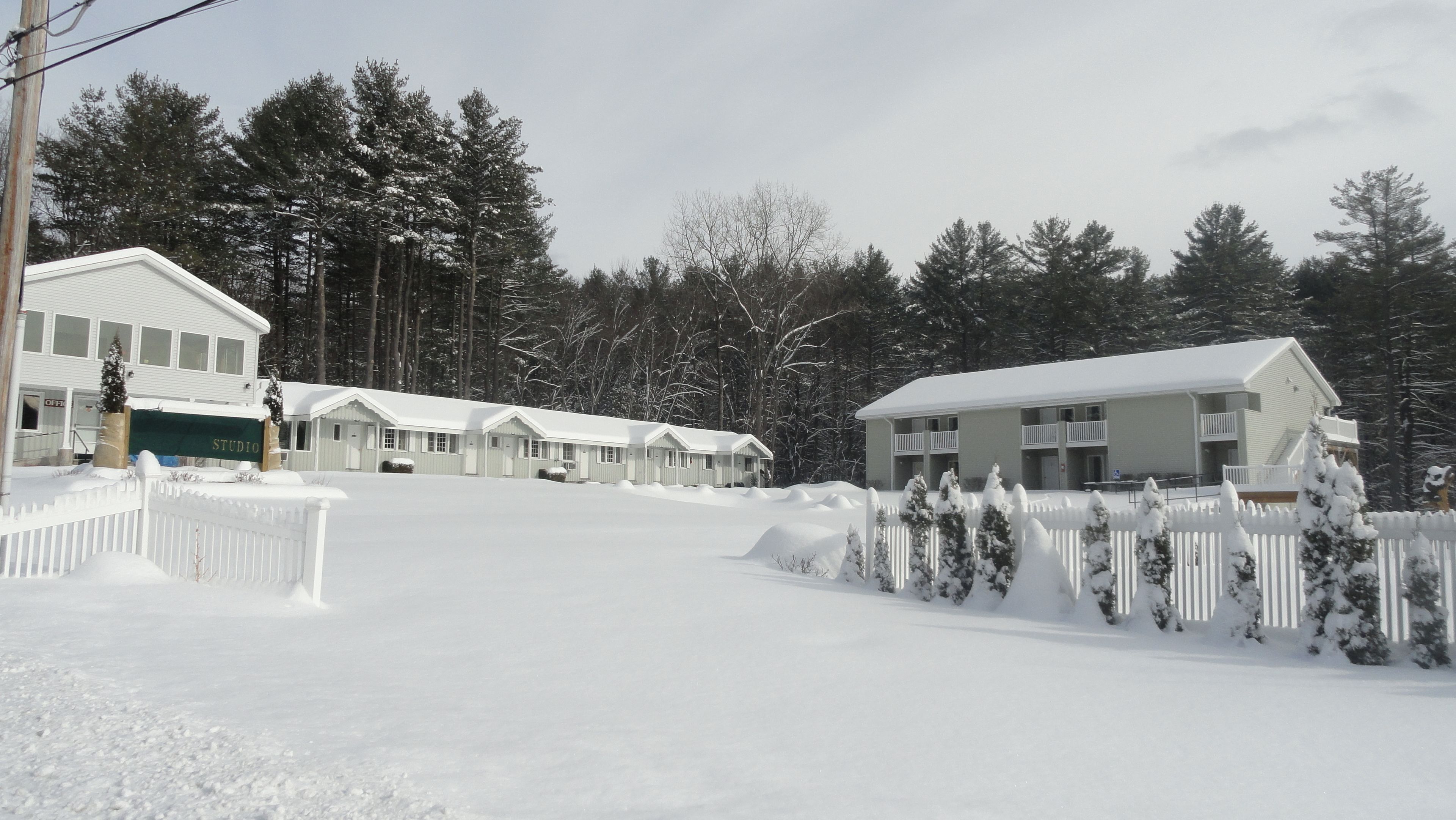 A snow-covered motel with multiple rooms, a fenced parking area, and a backdrop of trees.