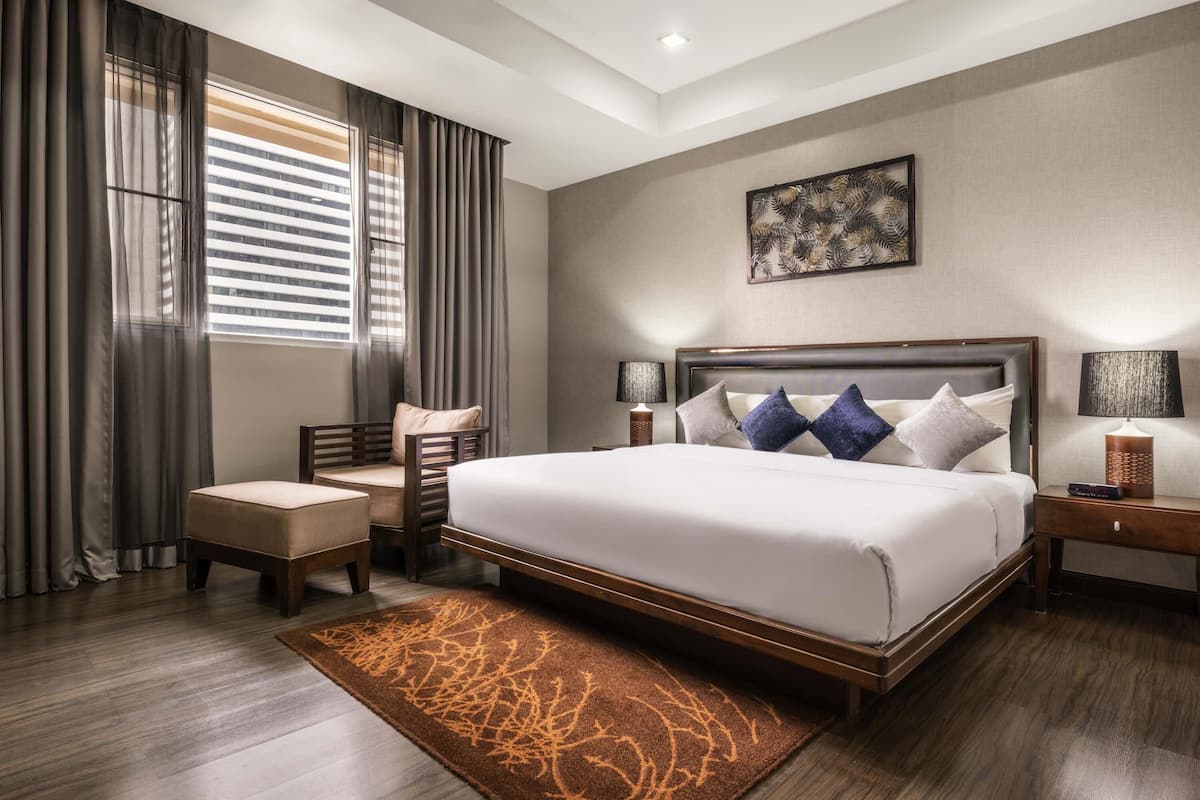 Hotelkamer met een groot bed, een stoel, een nachtkastje, een lamp, een vloerkleed en een raam met gordijnen.