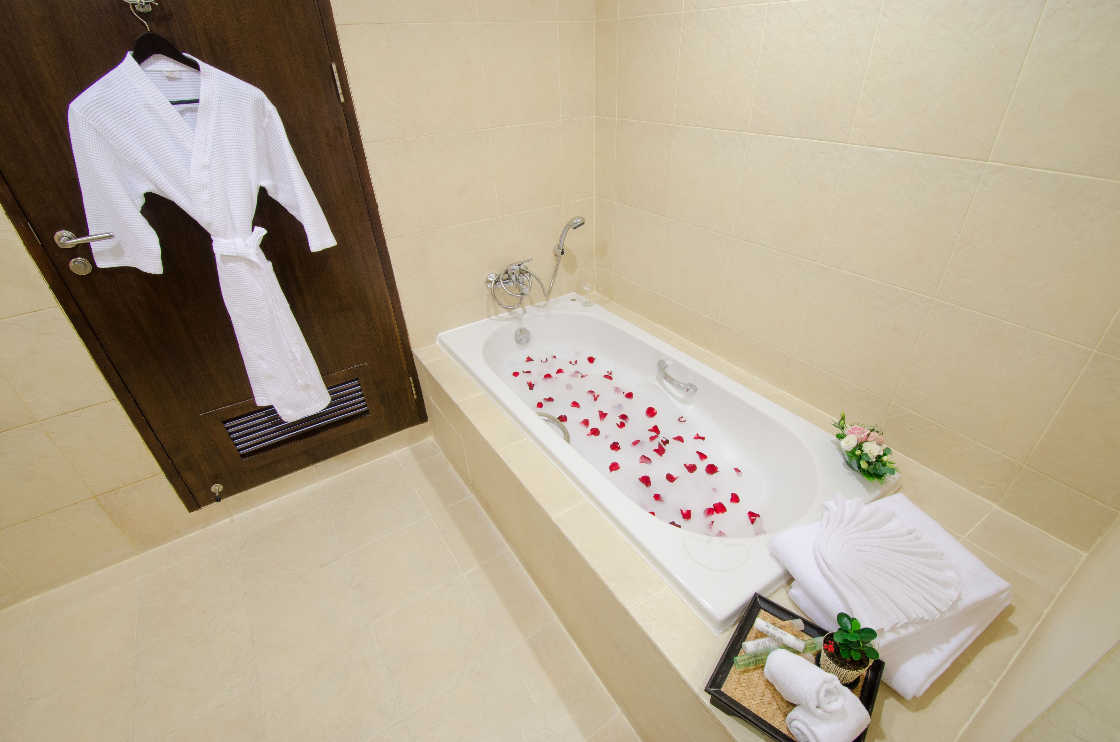 A bathroom with a bathtub filled with rose petals, a white robe hanging on a door, and a towel rack with folded towels.