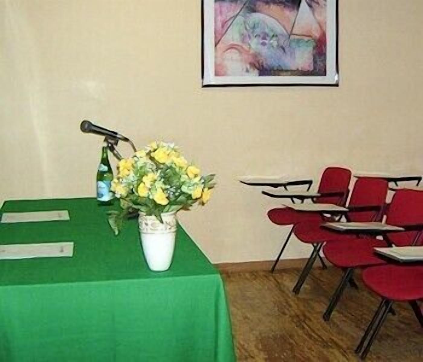 A conference room with a green table, a microphone, a vase with yellow flowers, and red chairs.