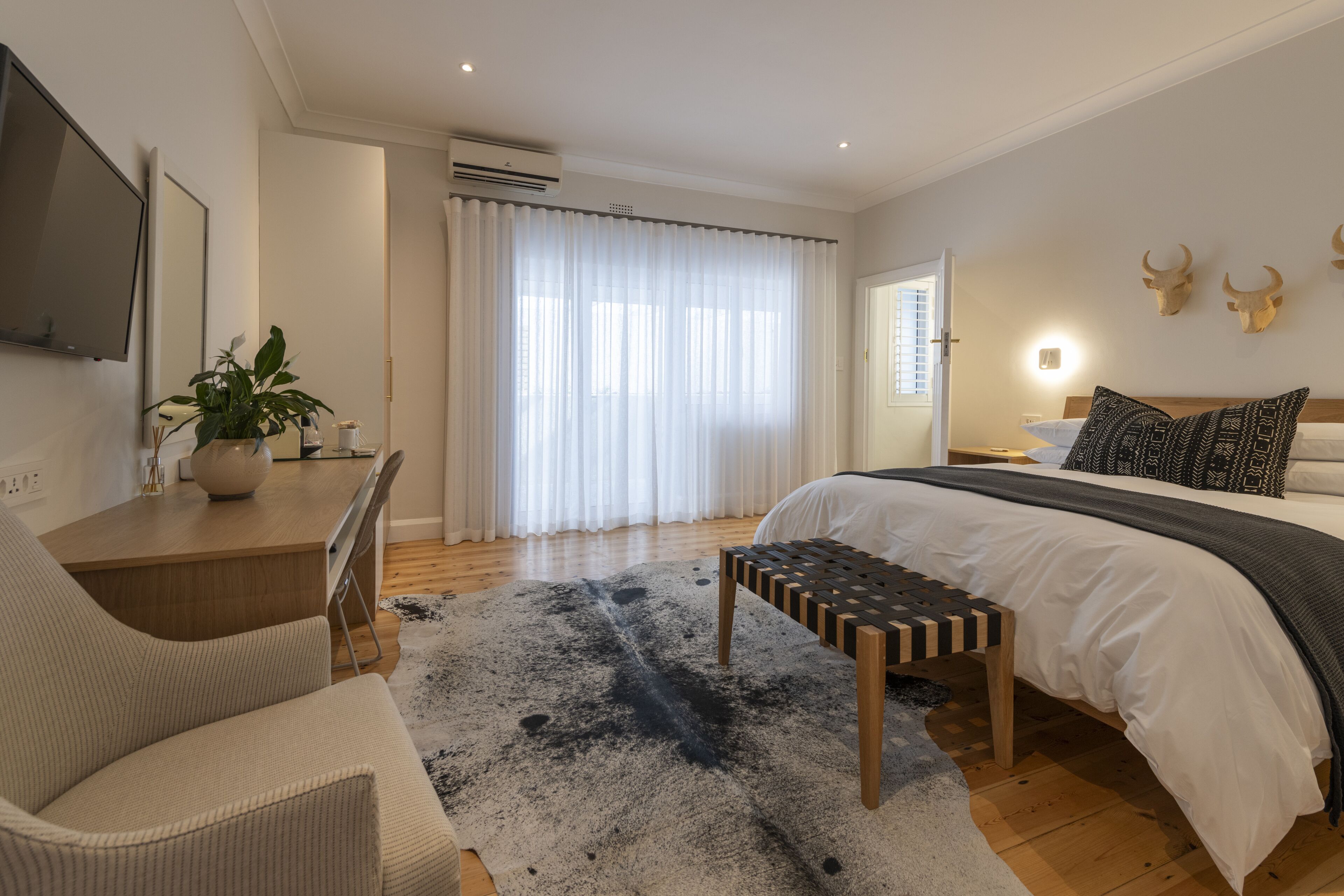 A modern bedroom with a large bed, a desk, a chair, and a large window with sheer curtains.