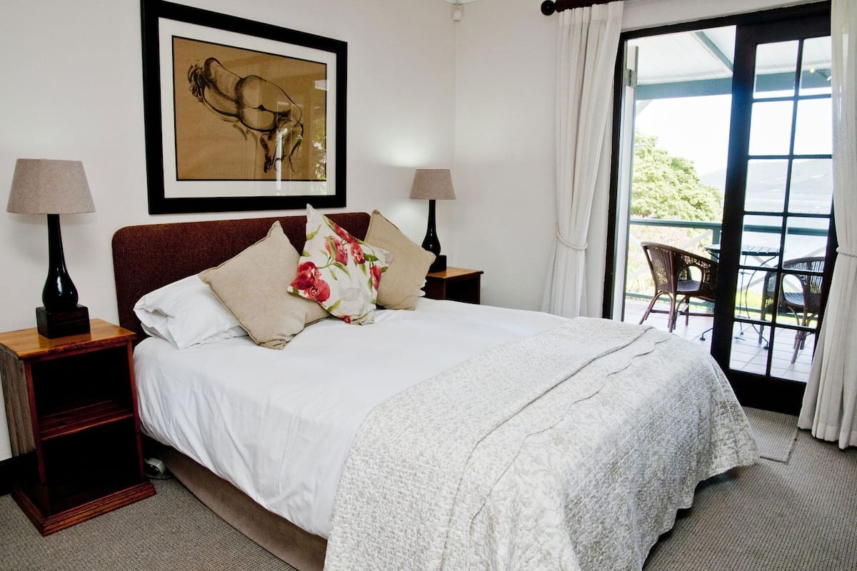 A neatly made bed with white linens, a floral pillow, and a framed artwork above it. There are two bedside tables with lamps, and a view of an outdoor patio through a glass door.
