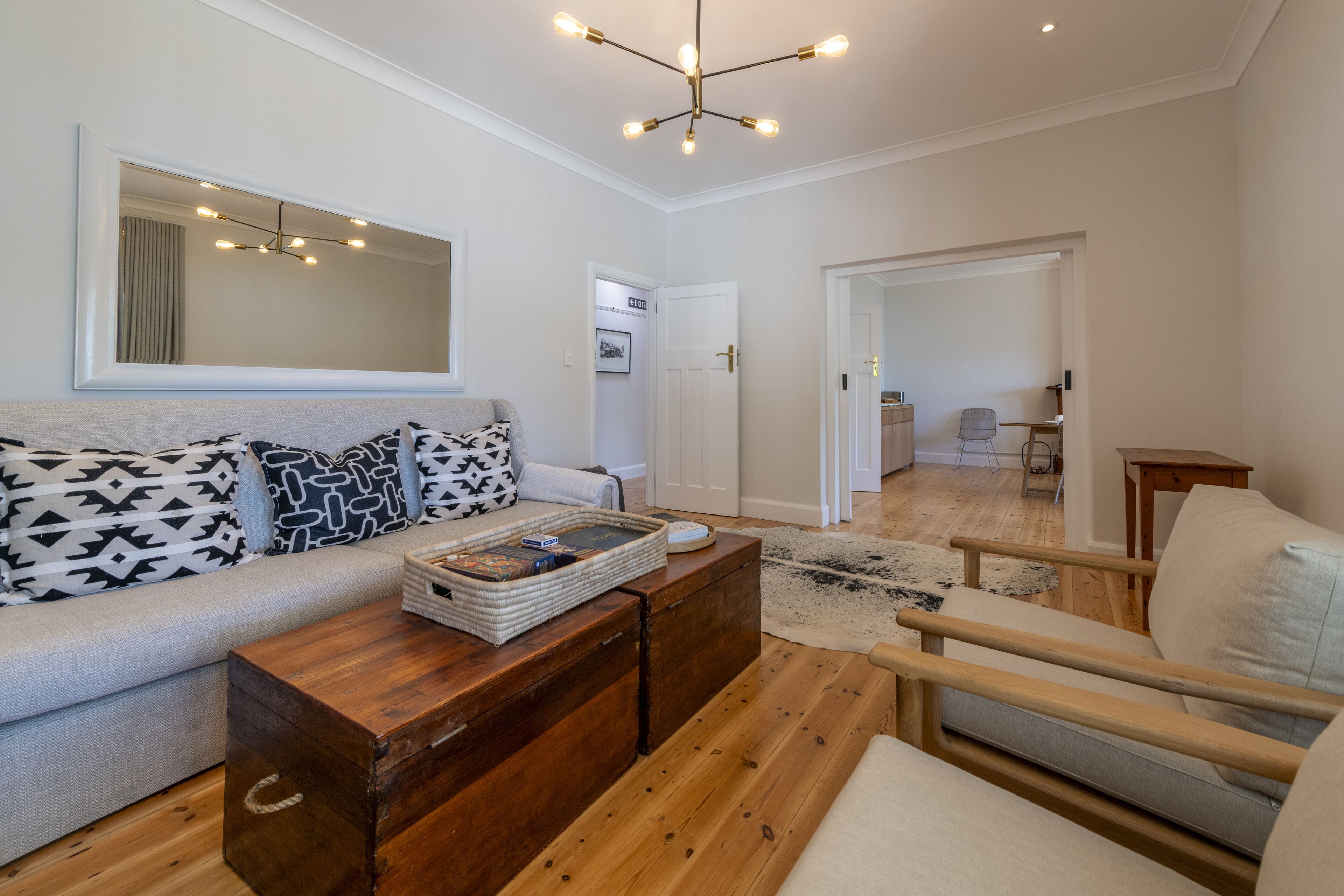 A modern living room with a sofa, coffee table, and a wooden chest. There is a mirror on the wall and a door leading to another room.