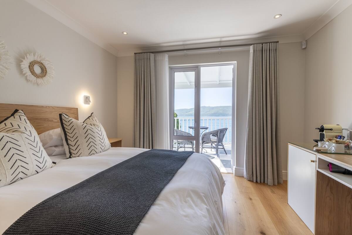 A hotel room with a large bed, a desk with a printer, and a balcony with a view of the sea.