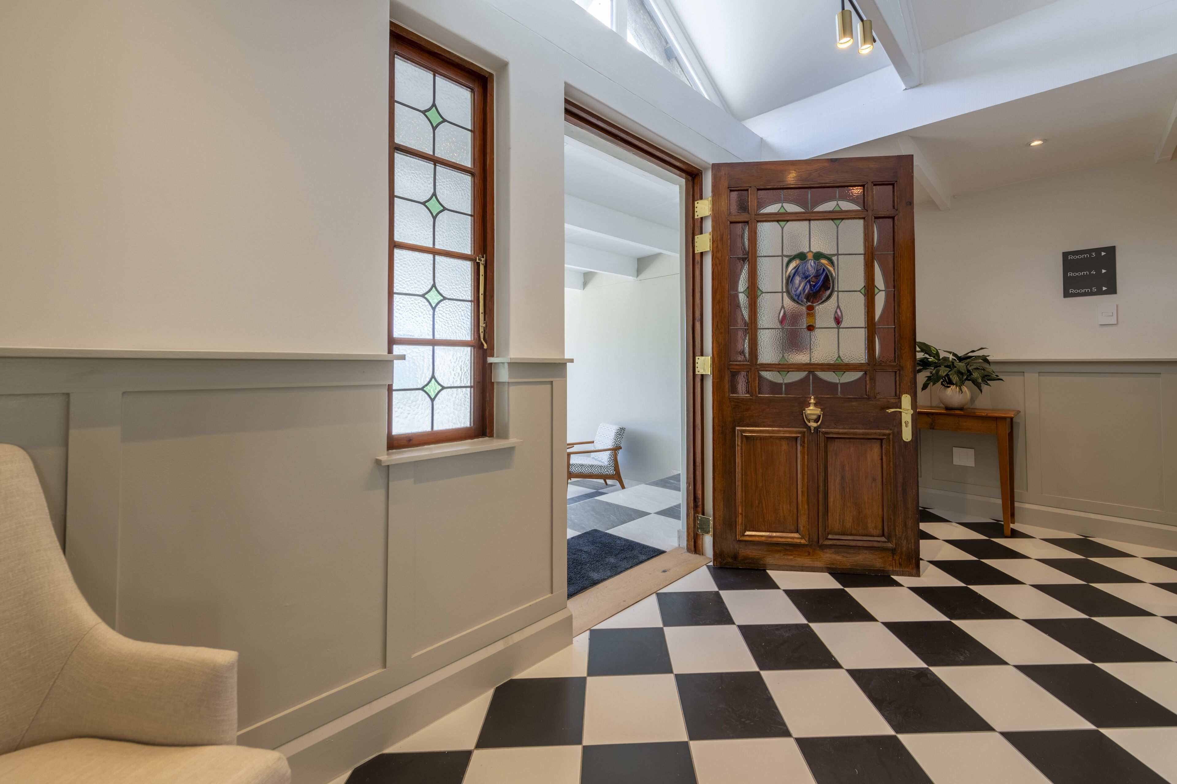 A hotel lobby with a checkered floor, a wooden door with glass panels, a beige armchair, and a sign indicating room numbers.