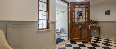 A hotel lobby with a checkered floor, a wooden door with glass panels, a beige armchair, and a sign indicating room numbers.