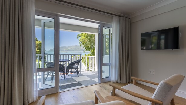 A modern room with a large sliding glass door leading to a balcony with a view of a lake and mountains. There is a flat-screen TV mounted on the wall and a wooden bench in the room. - Hide Away Guest House (Knysna)