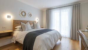 A modern bedroom with a wooden headboard, white bedding, and a grey throw blanket. There are two wall-mounted light fixtures and a large window with curtains. - Hide Away Guest House (Knysna)
