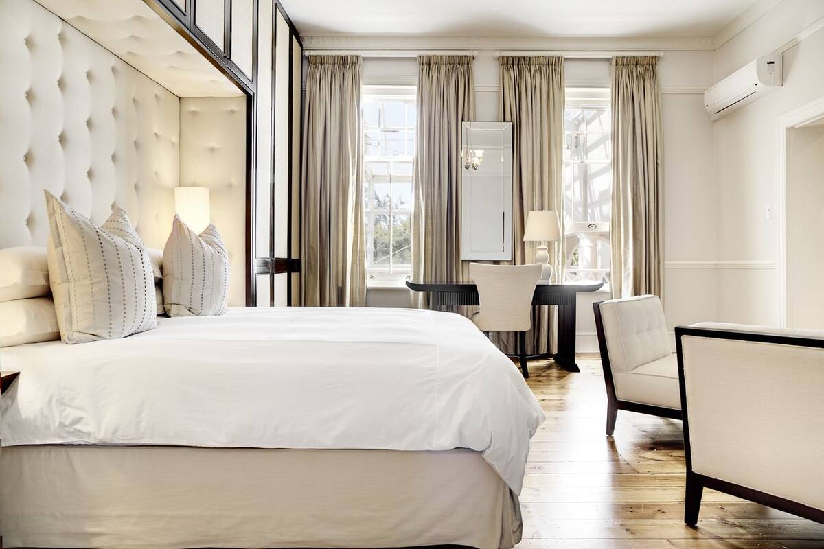 A hotel room with a large bed, a desk, and a chair. There is a window with curtains, and an air conditioning unit on the wall.