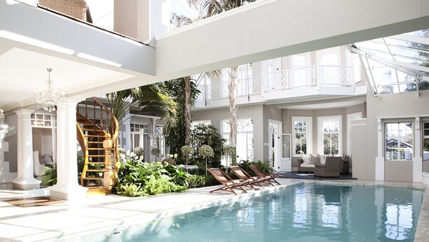 A spacious indoor pool area with a wooden staircase, lounge chairs, and a variety of plants.