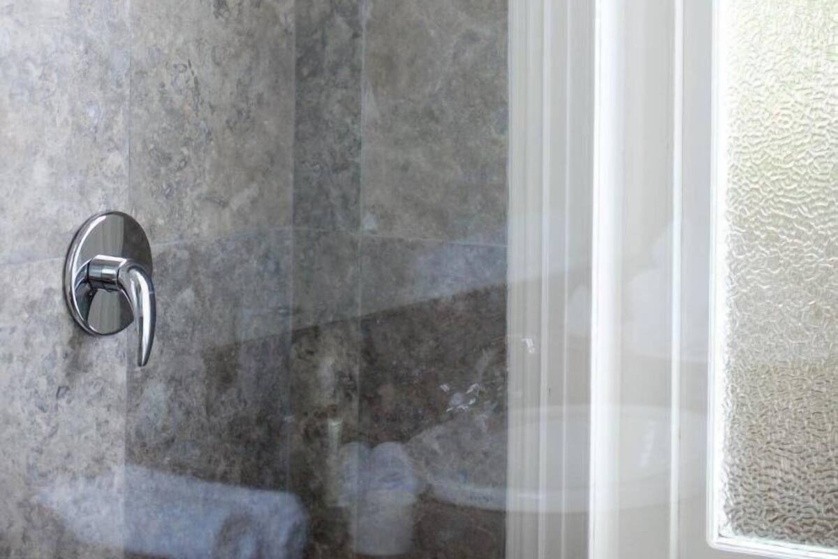 A modern shower with a glass shelf, marble walls, and a window with a textured view.