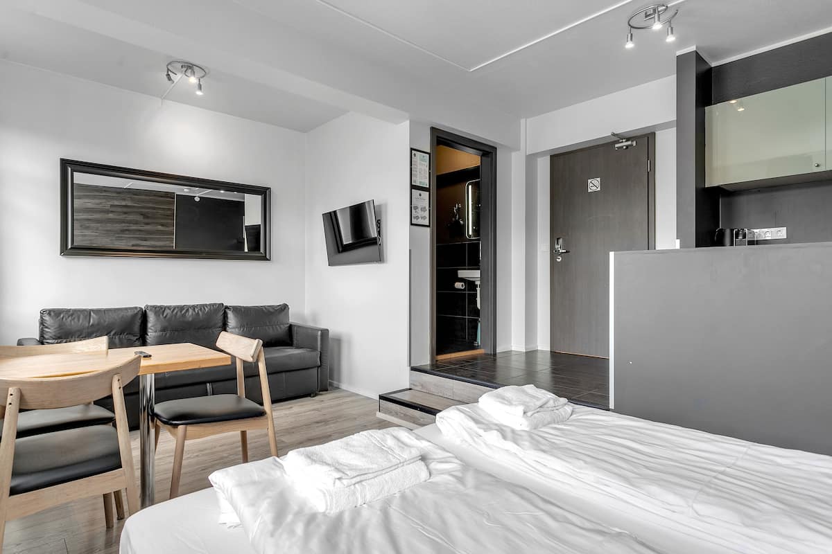 A modern hotel room with a bed, a desk with a chair, a sofa, a TV mounted on the wall, and a door leading to a bathroom.