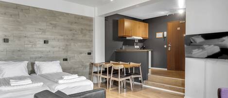 Une chambre d’hôtel moderne avec une tête de lit en bois, un canapé, une table à manger avec des chaises, un coin cuisine et un téléviseur.