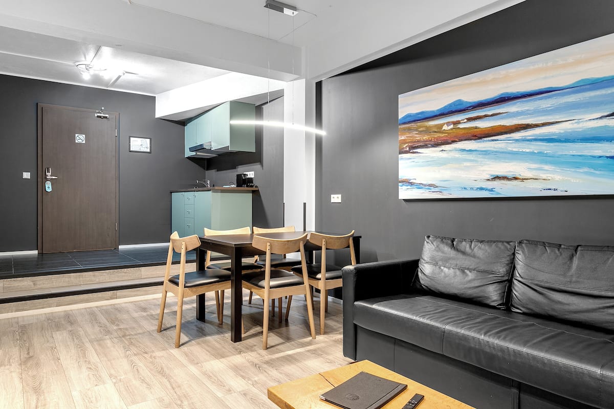 A modern living room with a black leather sofa, wooden dining table, and chairs. A painting is on the wall, and a ceiling fan is visible.