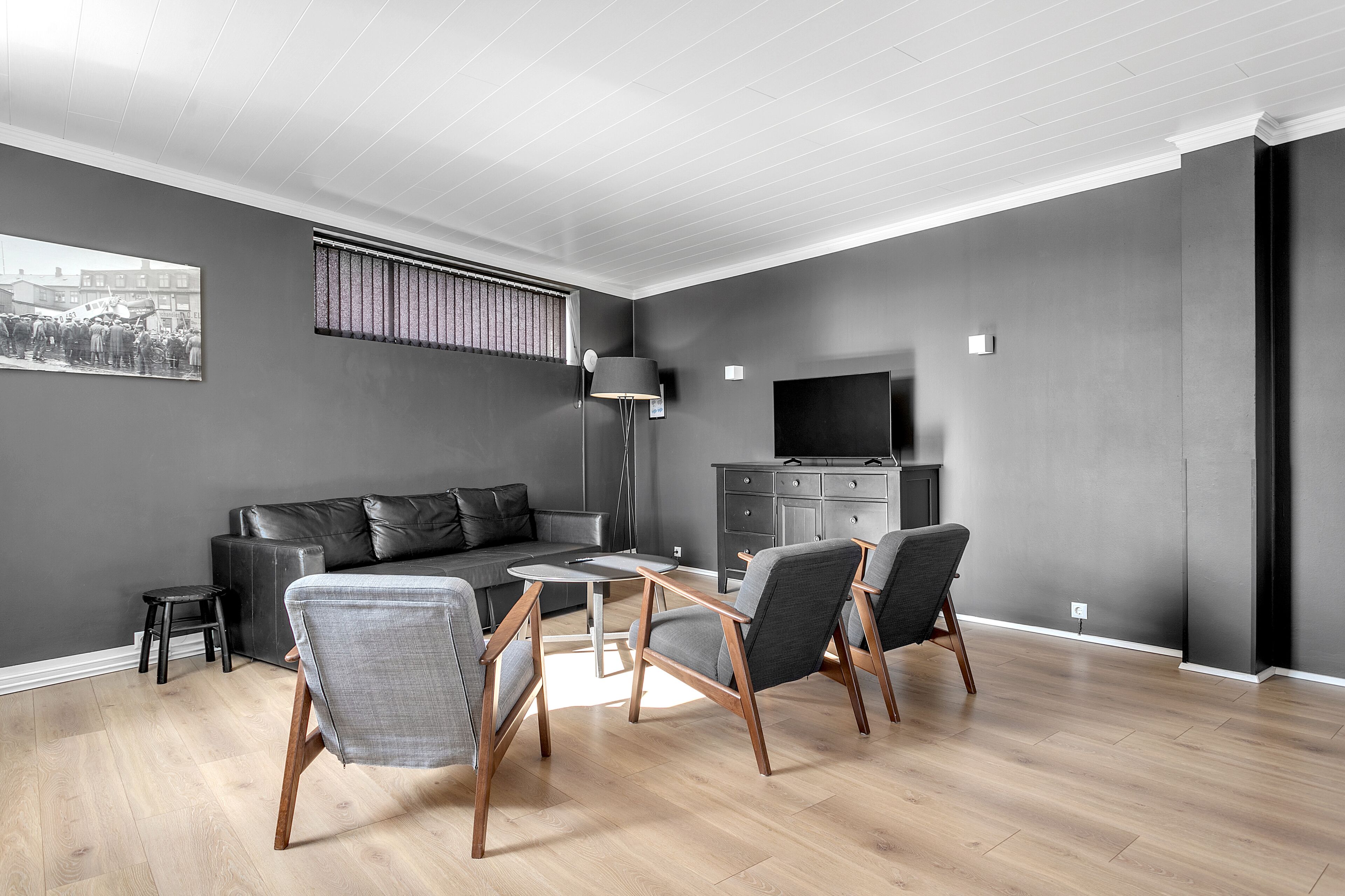 Una sala de estar moderna con un sofá gris, dos sillones grises, una mesita de centro, una lámpara de pie, un televisor y una pequeña mesita auxiliar.