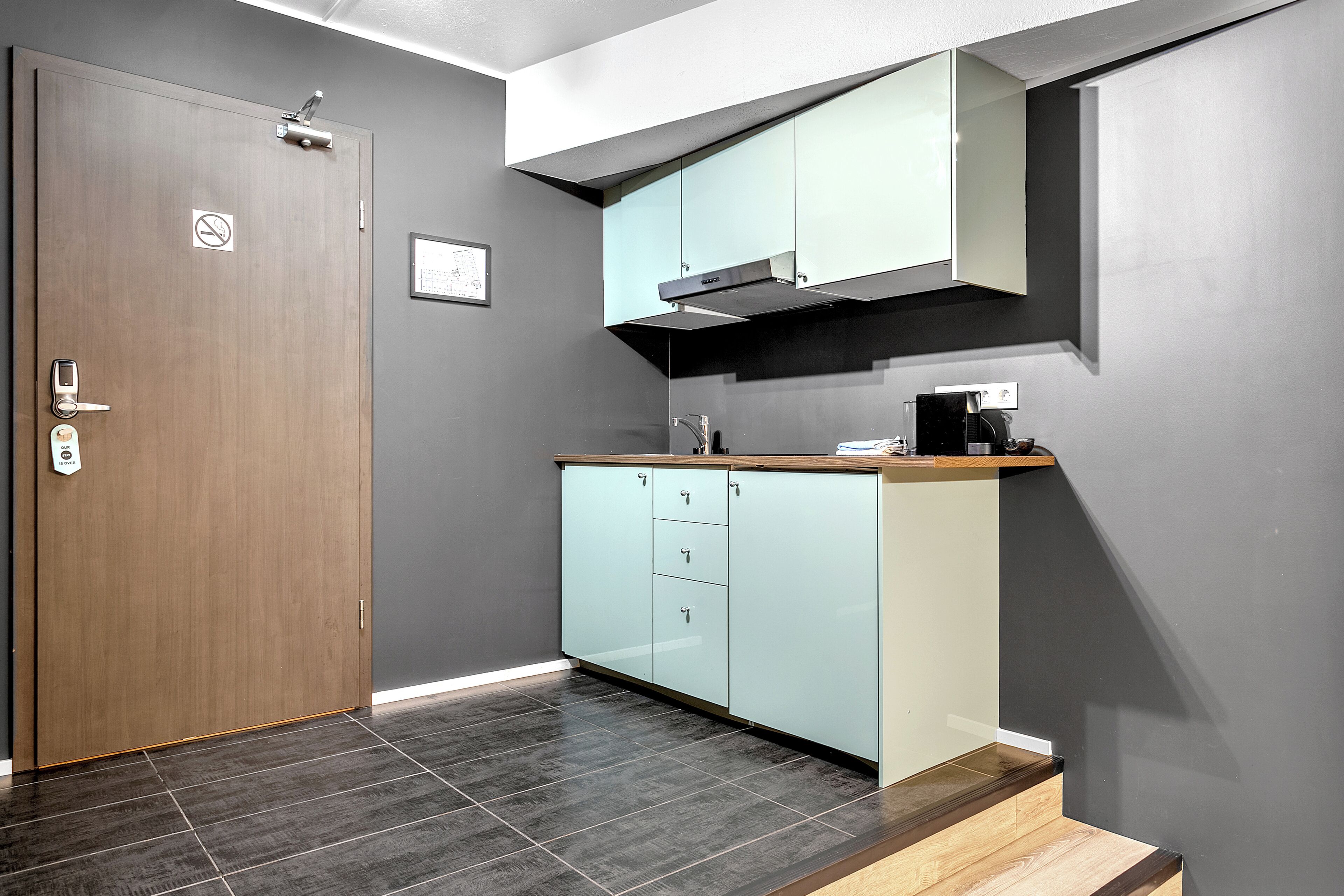 A modern kitchen with a wooden door, light blue cabinets, and a dark tiled floor.