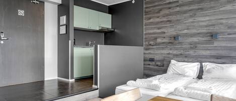 A modern hotel room with a wooden floor, a sofa bed, a wooden dining table, and a kitchen area with a built-in oven and cabinets.