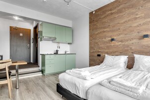 Un dormitorio moderno con cabecera de madera, cama con ropa de cama blanca y una cocina verde con pileta y alacenas.