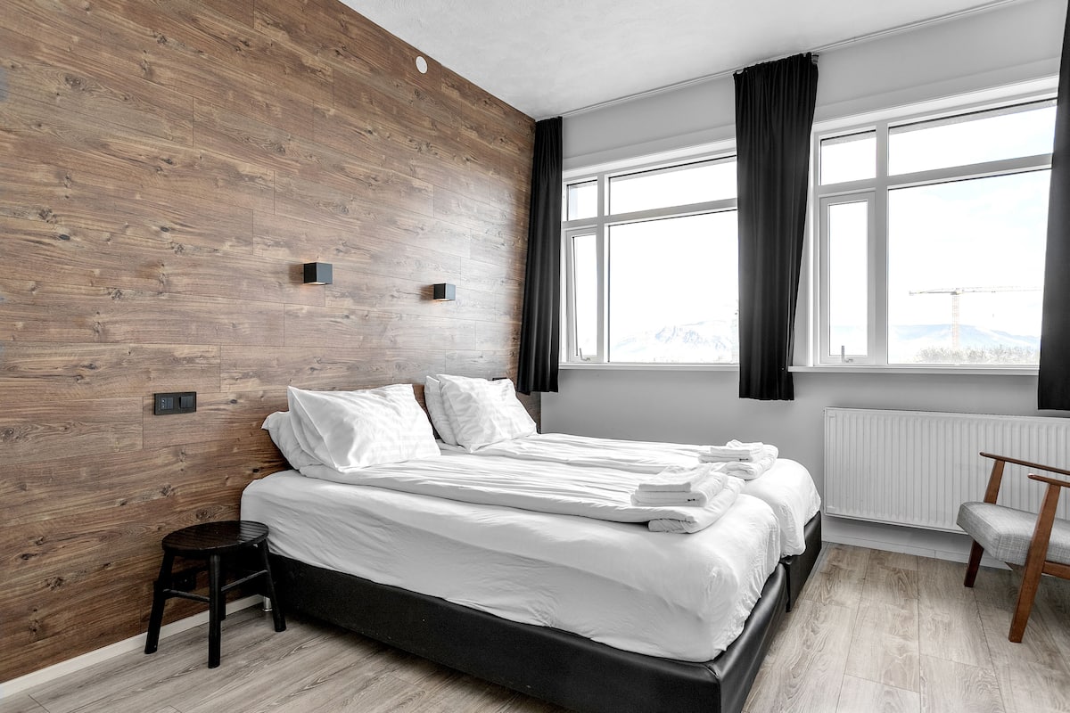 A modern bedroom with a wooden wall, a large window with curtains, a bed with white linens, and a small wooden stool.