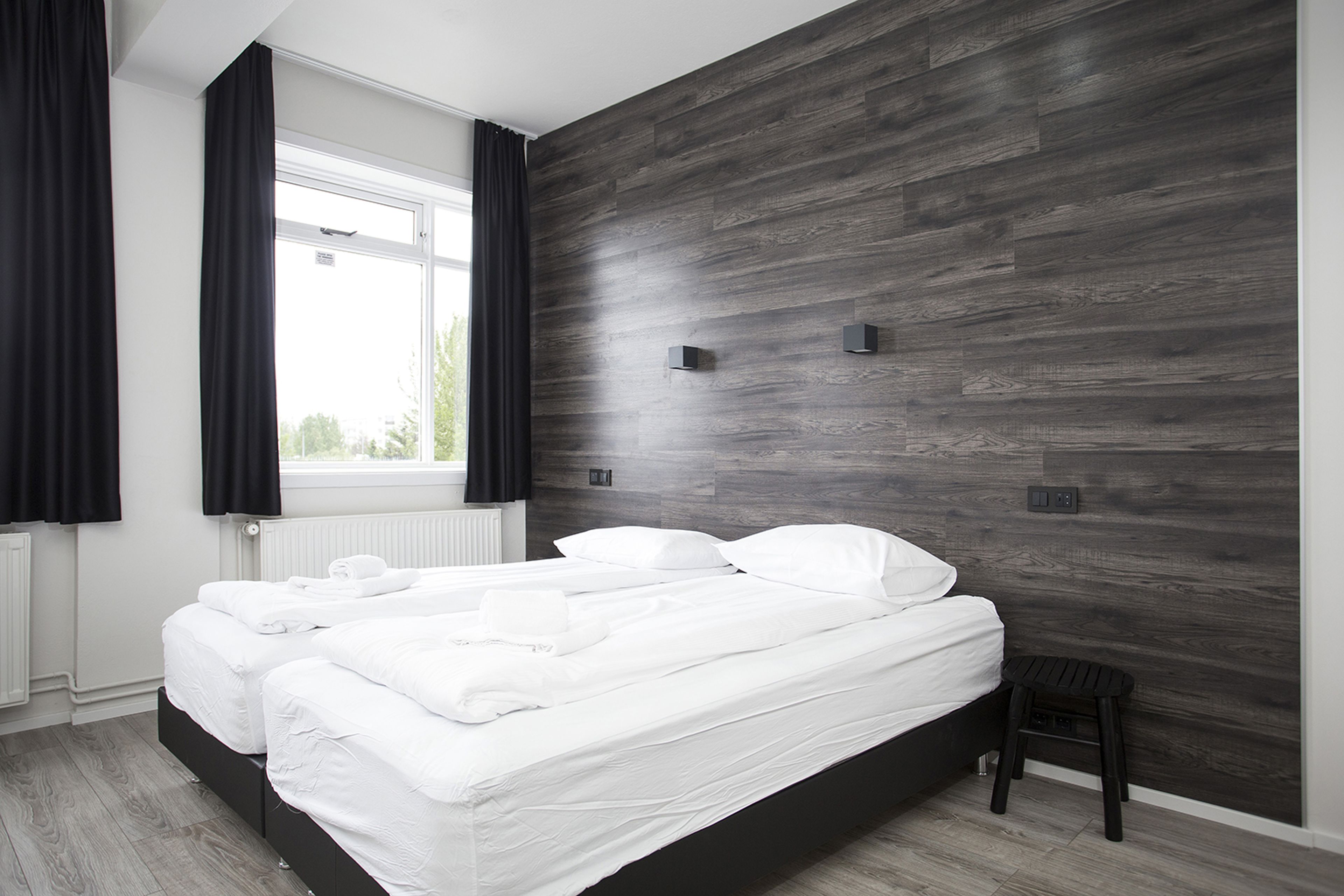 A modern bedroom with a large bed, a small stool, and a window with curtains.