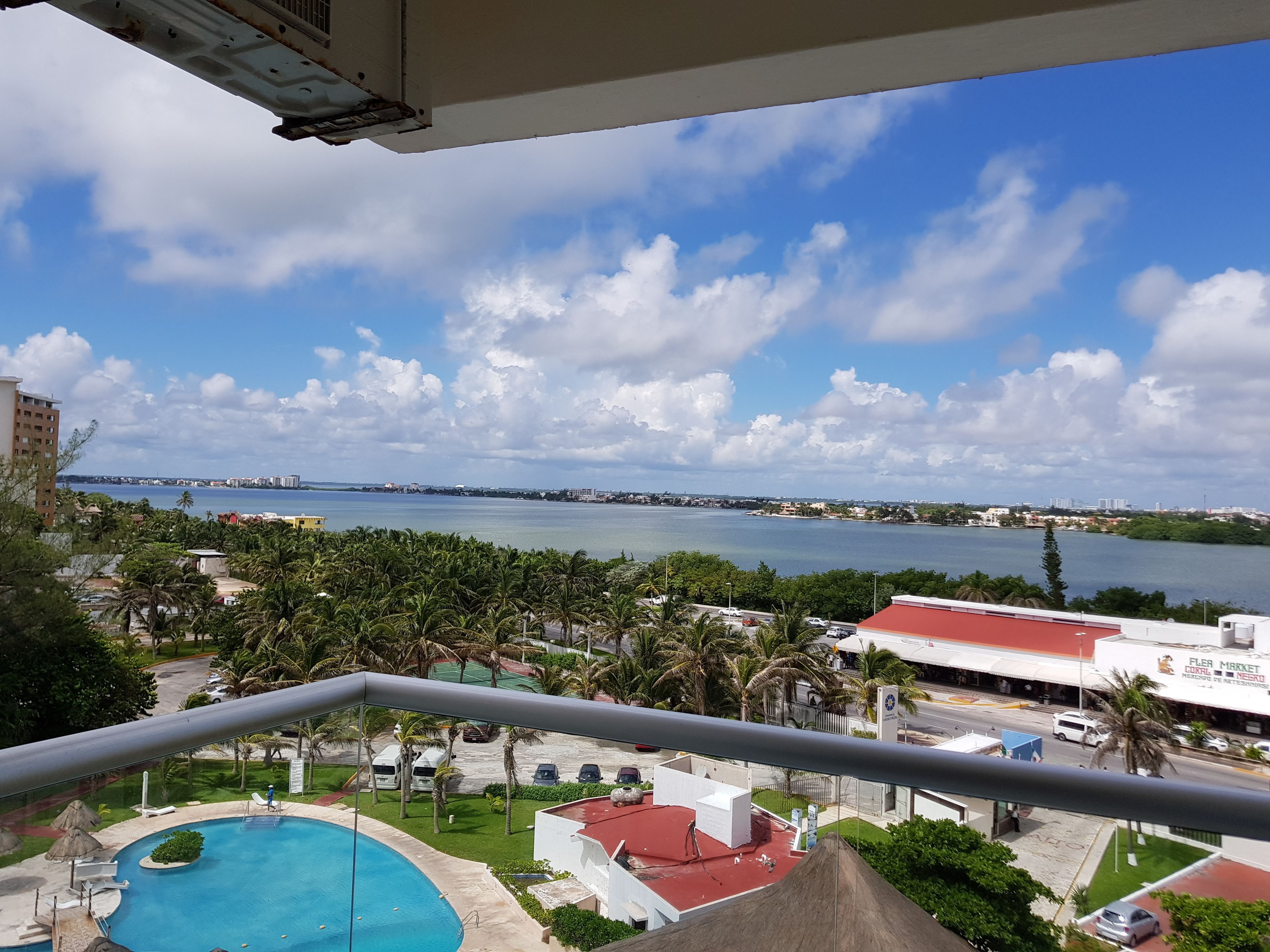 A view from a balcony overlooking a swimming pool, a body of water, and a distant cityscape.
