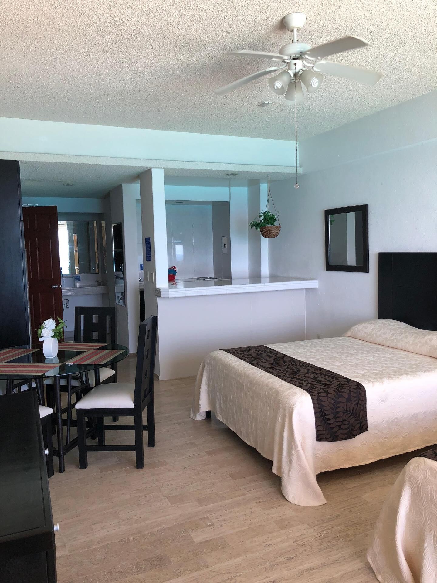 Una habitación de hotel con cama, una mesa de comedor con sillas, un ventilador de techo y una planta colgante.