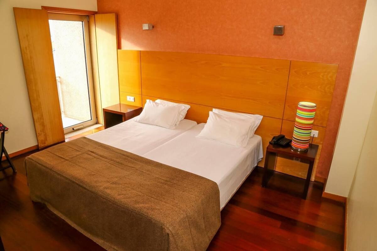 A neatly made bed with white linens and a brown throw blanket, a wooden headboard, a bedside table with a lamp and a phone, and a window with a view of the outside.