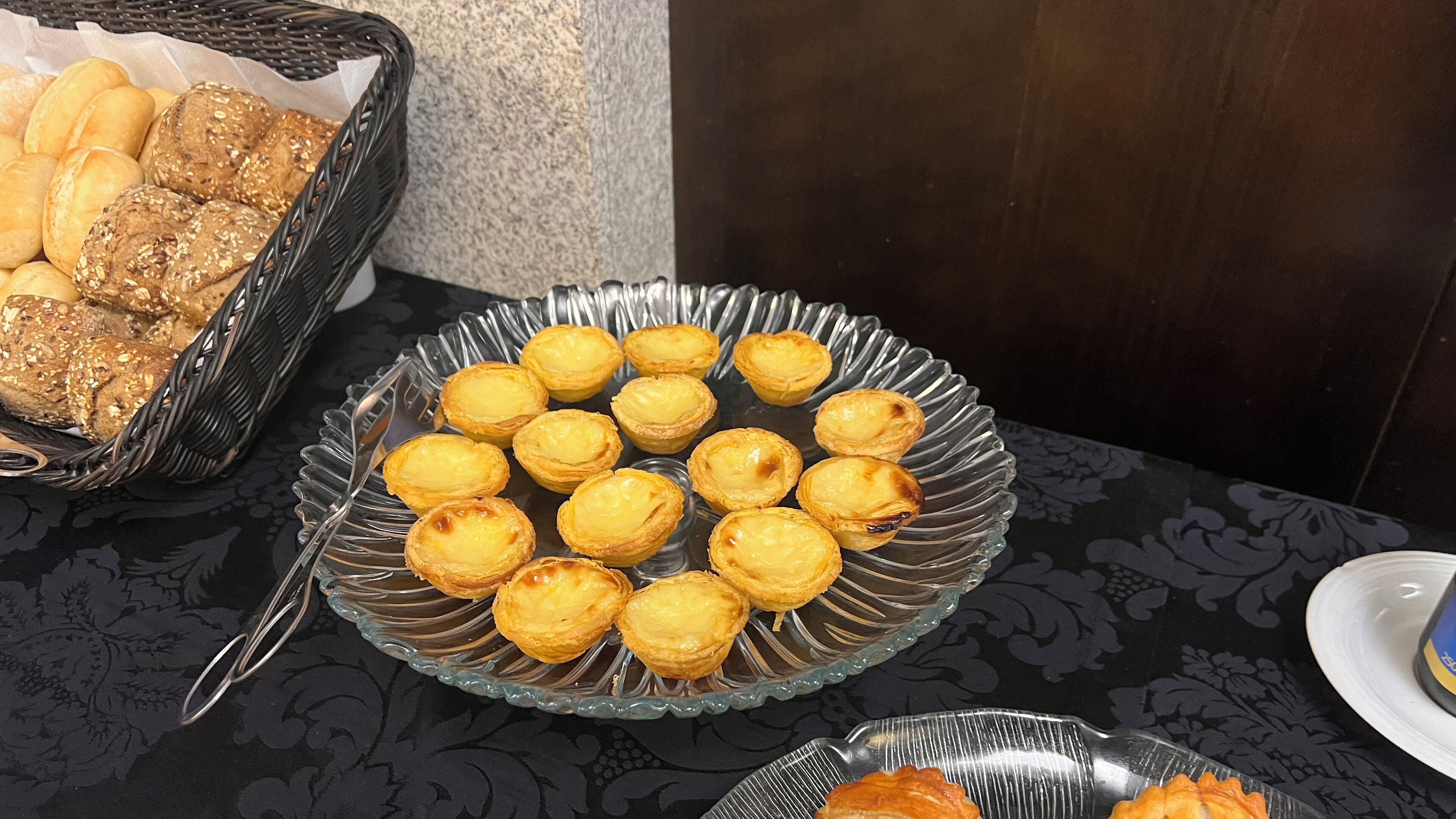 Une table sur laquelle se trouvent une assiette en verre remplie de tartelettes aux œufs et un panier de petits pains.