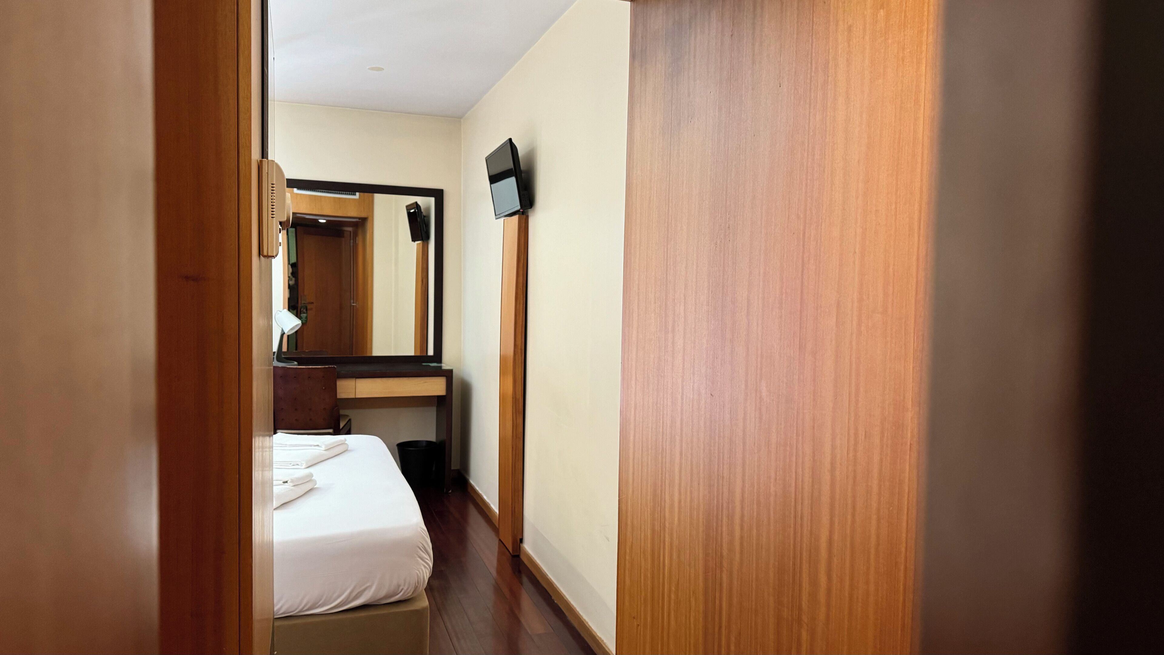 A narrow hotel hallway with a bed, a desk, a mirror, and a flat-screen TV.