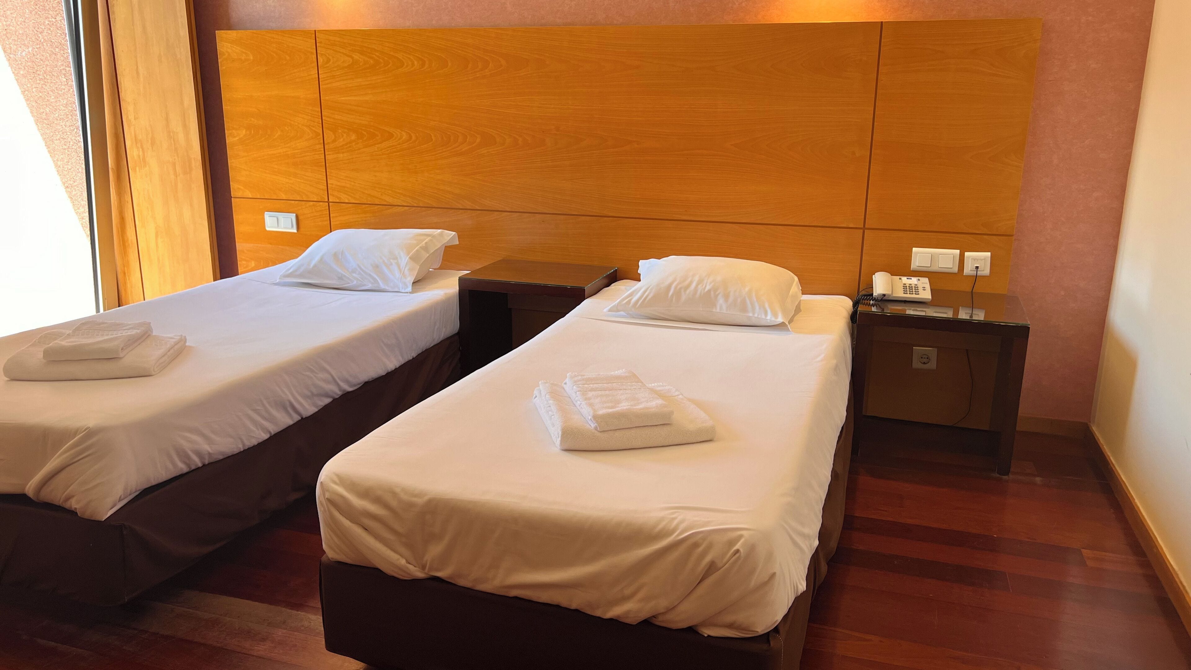 Two single beds with white linens and towels, a wooden headboard, a bedside table with a phone, and a wall socket.