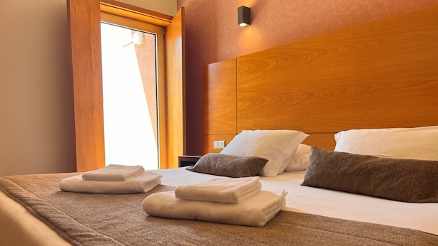 A neatly made bed with folded towels and pillows, a wooden headboard, and a window with curtains. - Flag Hotel Barcelos (Barcelos)