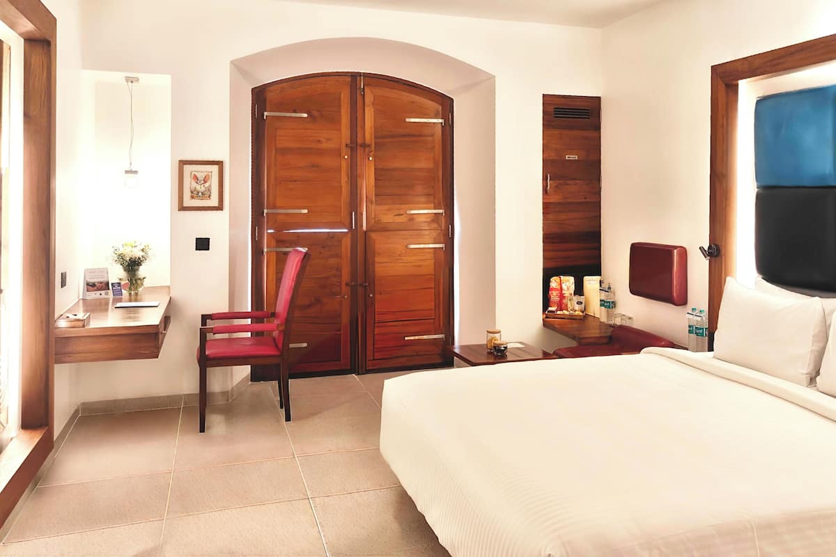 A hotel room with a bed, a desk with a chair, a wardrobe, and a small table with items.