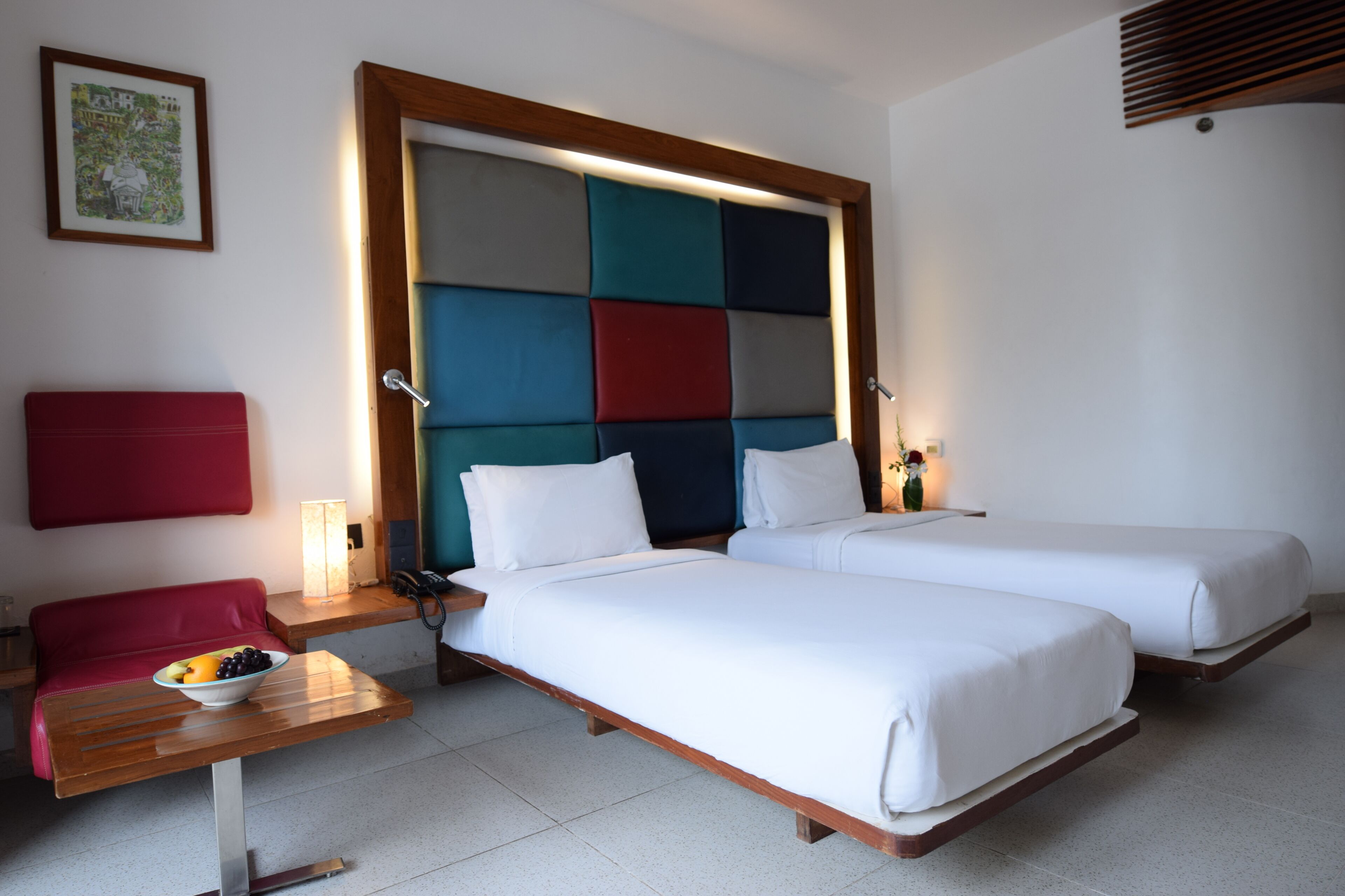 Habitación de hotel con dos camas, un sofá rojo, una mesa de madera con un frutero y un cabecero colorido.