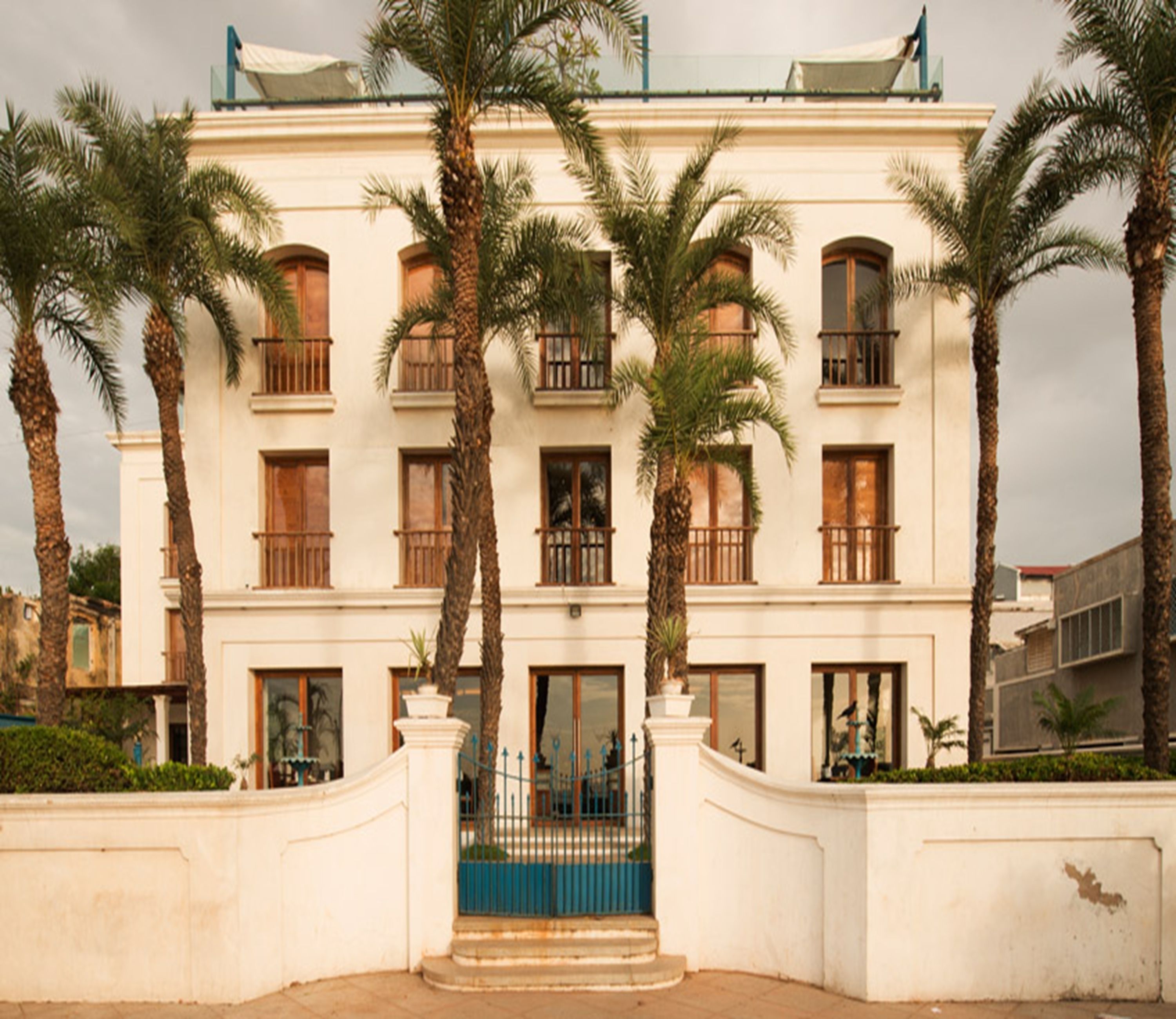 Un edificio de varios pisos con fachada simétrica, que presenta una entrada central con un portón azul, flanqueada por palmeras y una pared blanca con balaustrada decorativa.