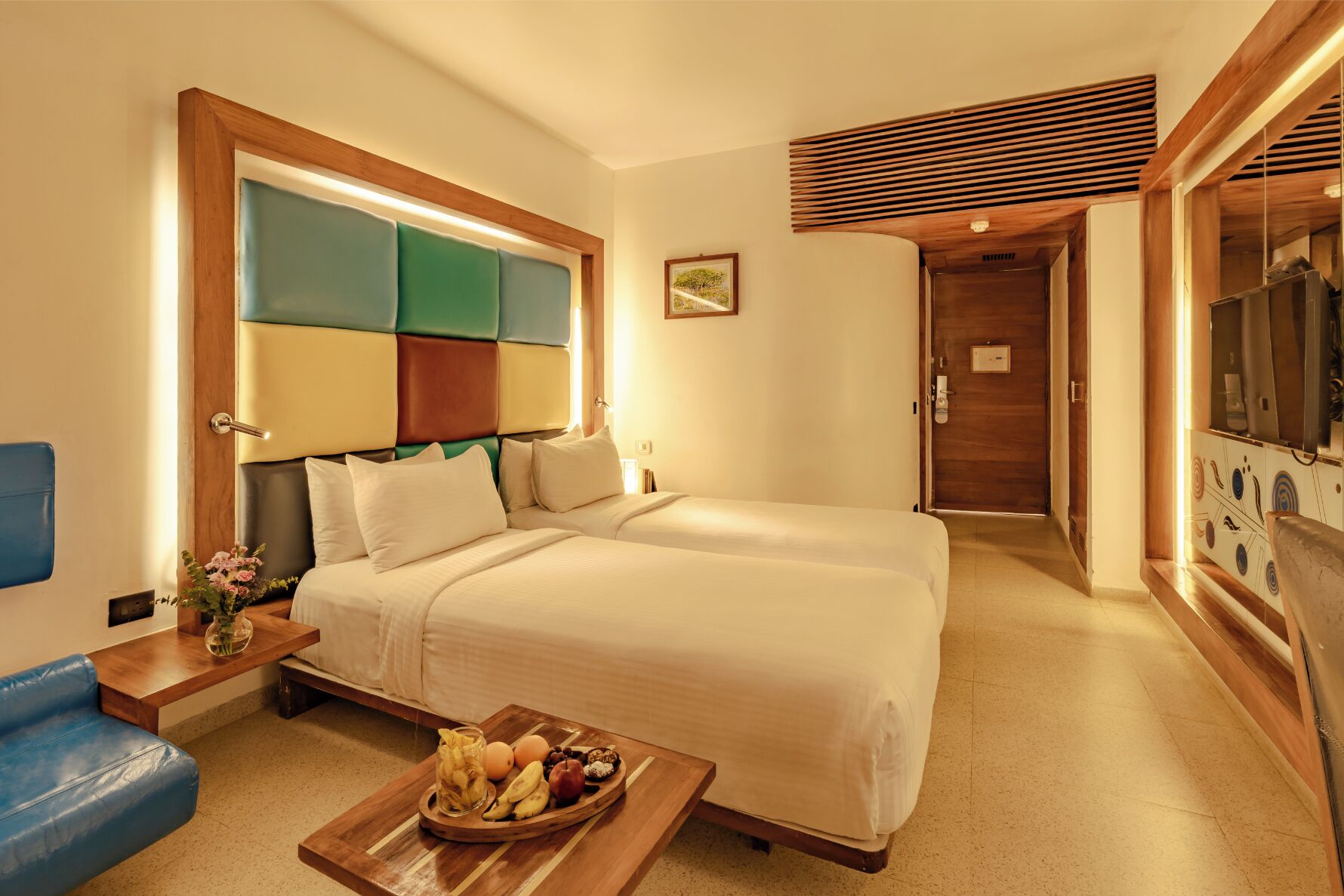 A hotel room with a large bed, a small table with snacks, a blue chair, and a wooden headboard.