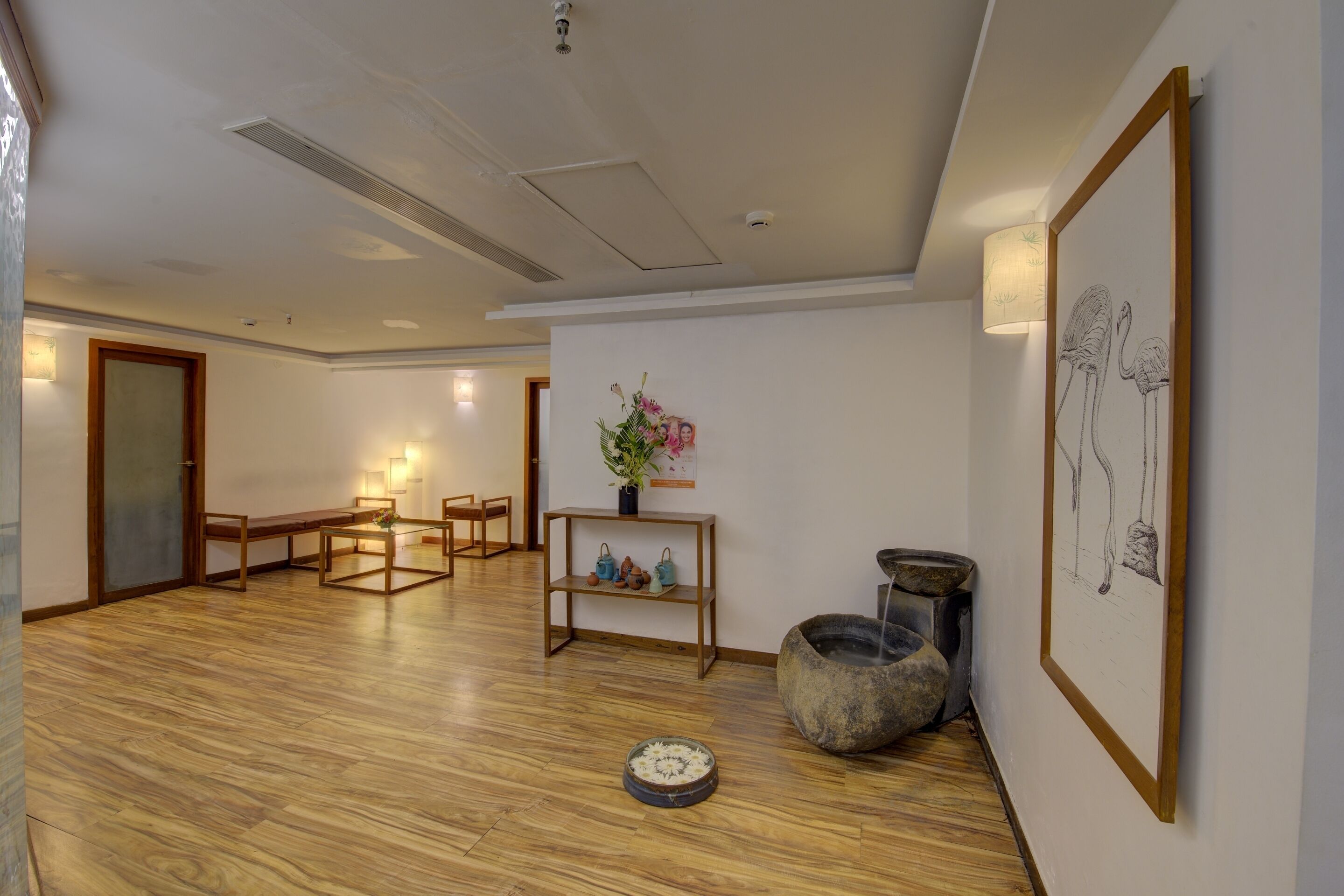 A spacious room with a wooden floor, a dining table with chairs, a small table with a vase and decorative items, and a framed artwork on the wall.