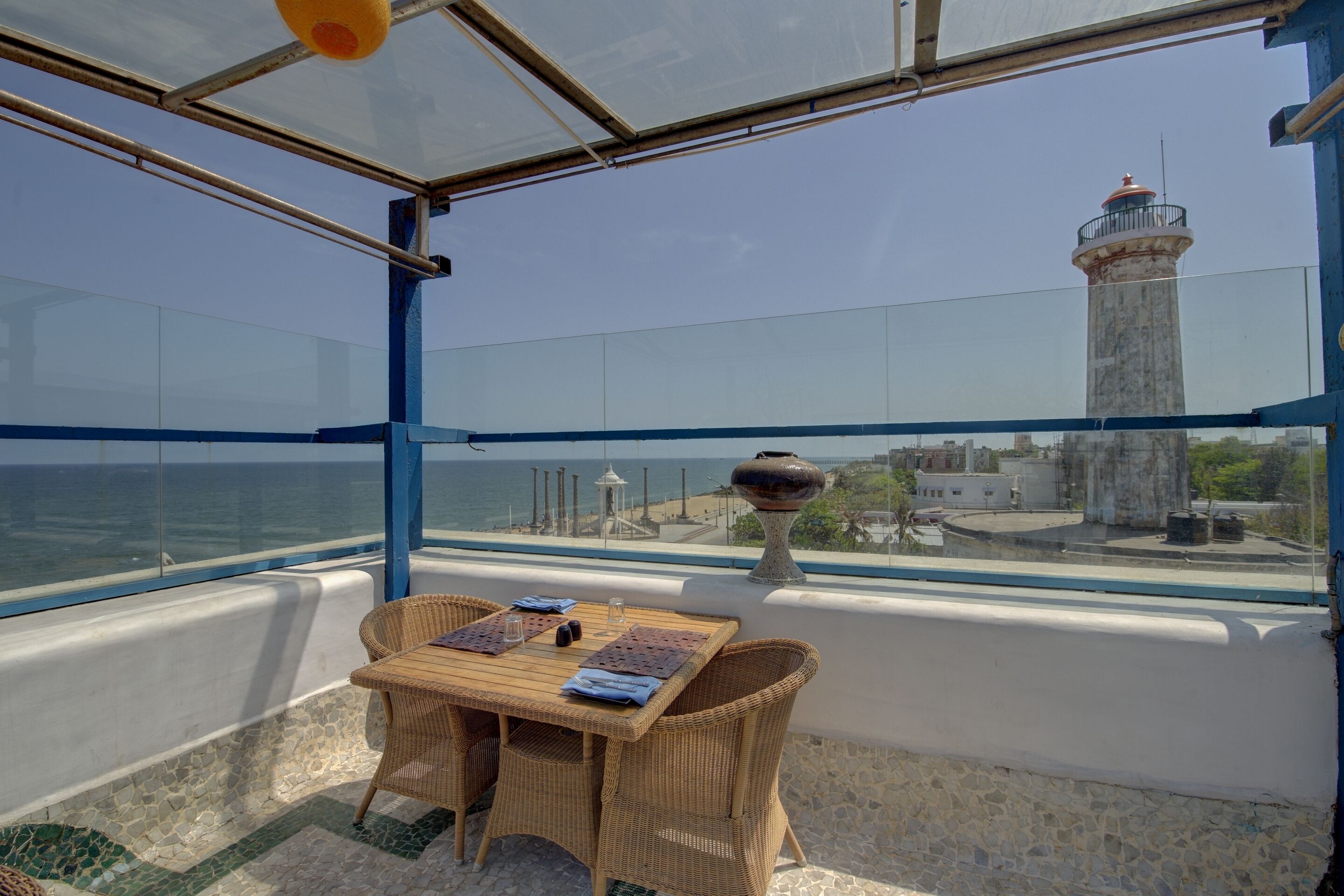 A covered terrace with a table and chairs, overlooking the sea and a lighthouse.