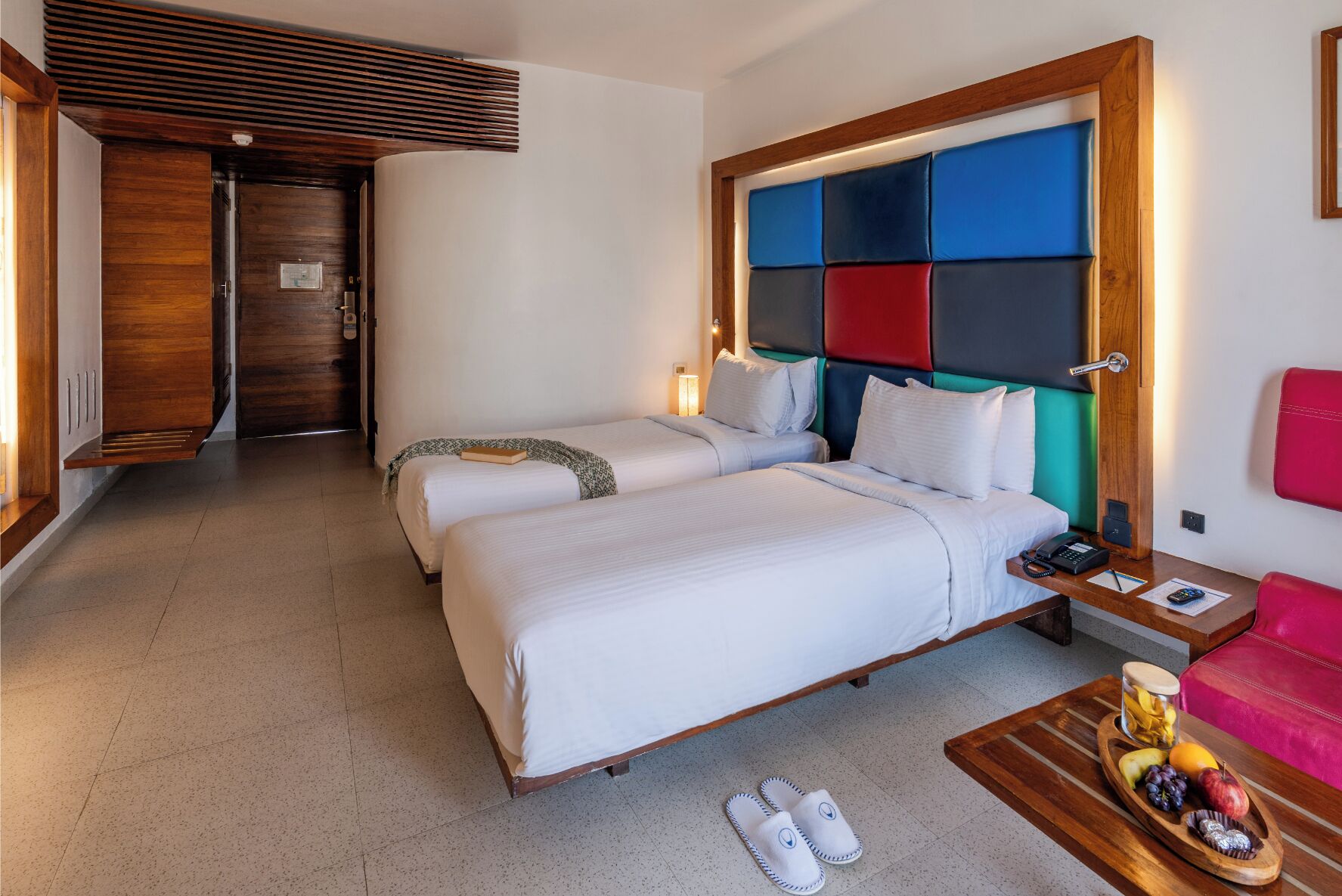 Ein Hotelzimmer mit zwei Betten, einem hölzernen Kopfteil, einem Schreibtisch mit Telefon, einem roten Sessel, Hausschuhen und einer Obstplatte.