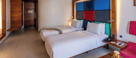 A hotel room with two beds, a wooden headboard, a desk with a phone, a red chair, slippers, and a fruit tray.