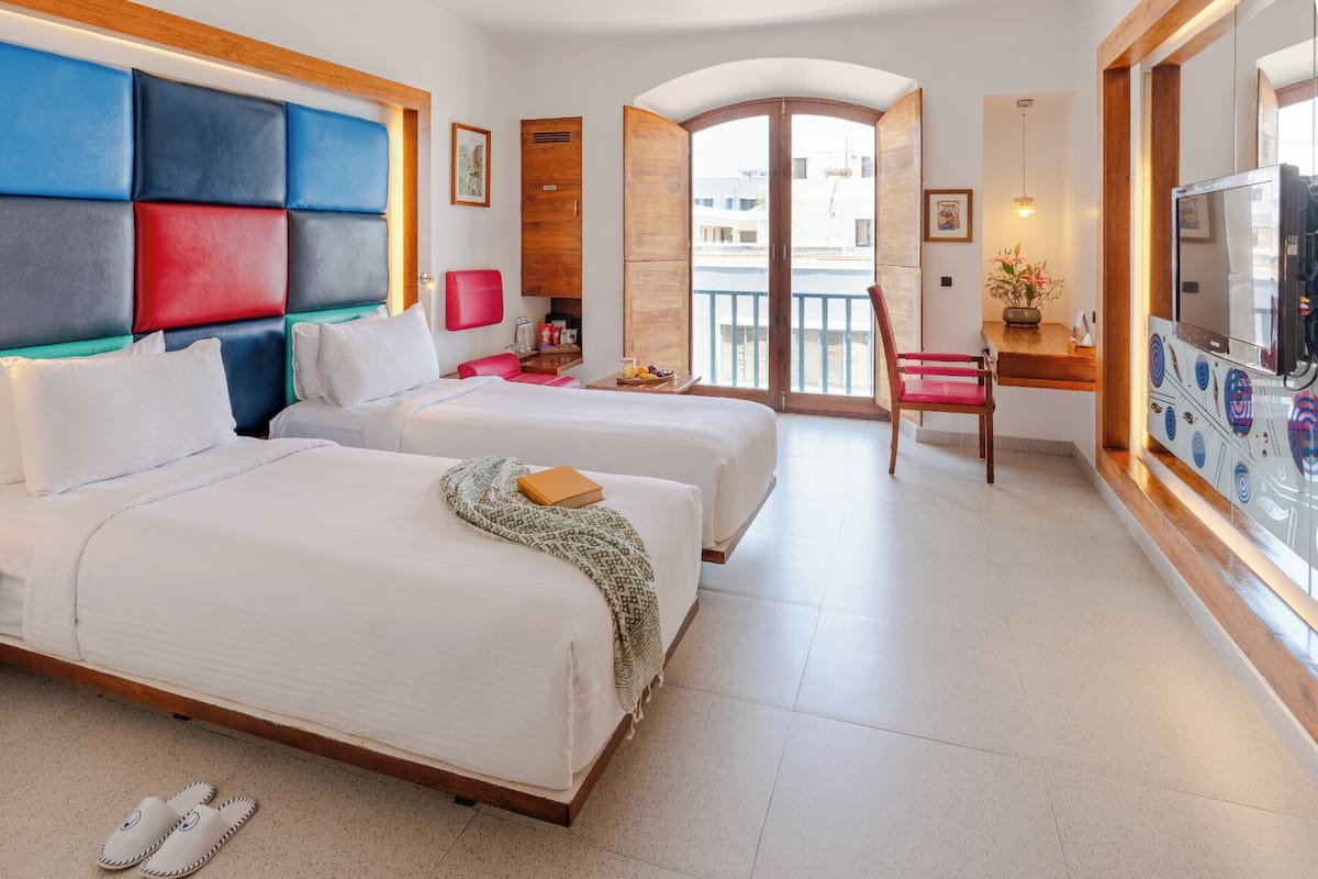 A hotel room with two beds, a desk, a chair, a TV, and a balcony with a view.