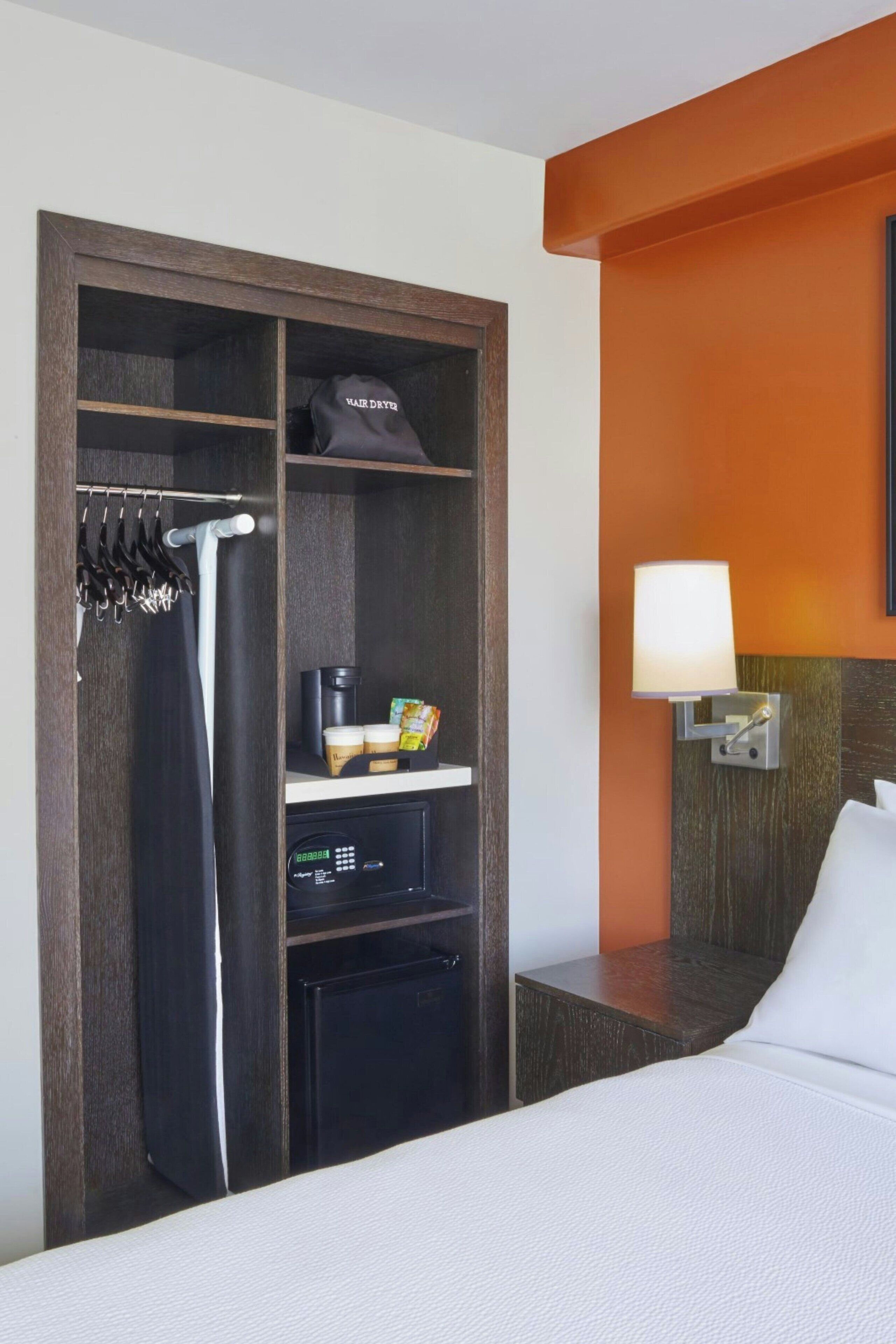 A hotel room with a bed, a wardrobe, a nightstand with a lamp, and a small desk with a coffee maker and snacks.
