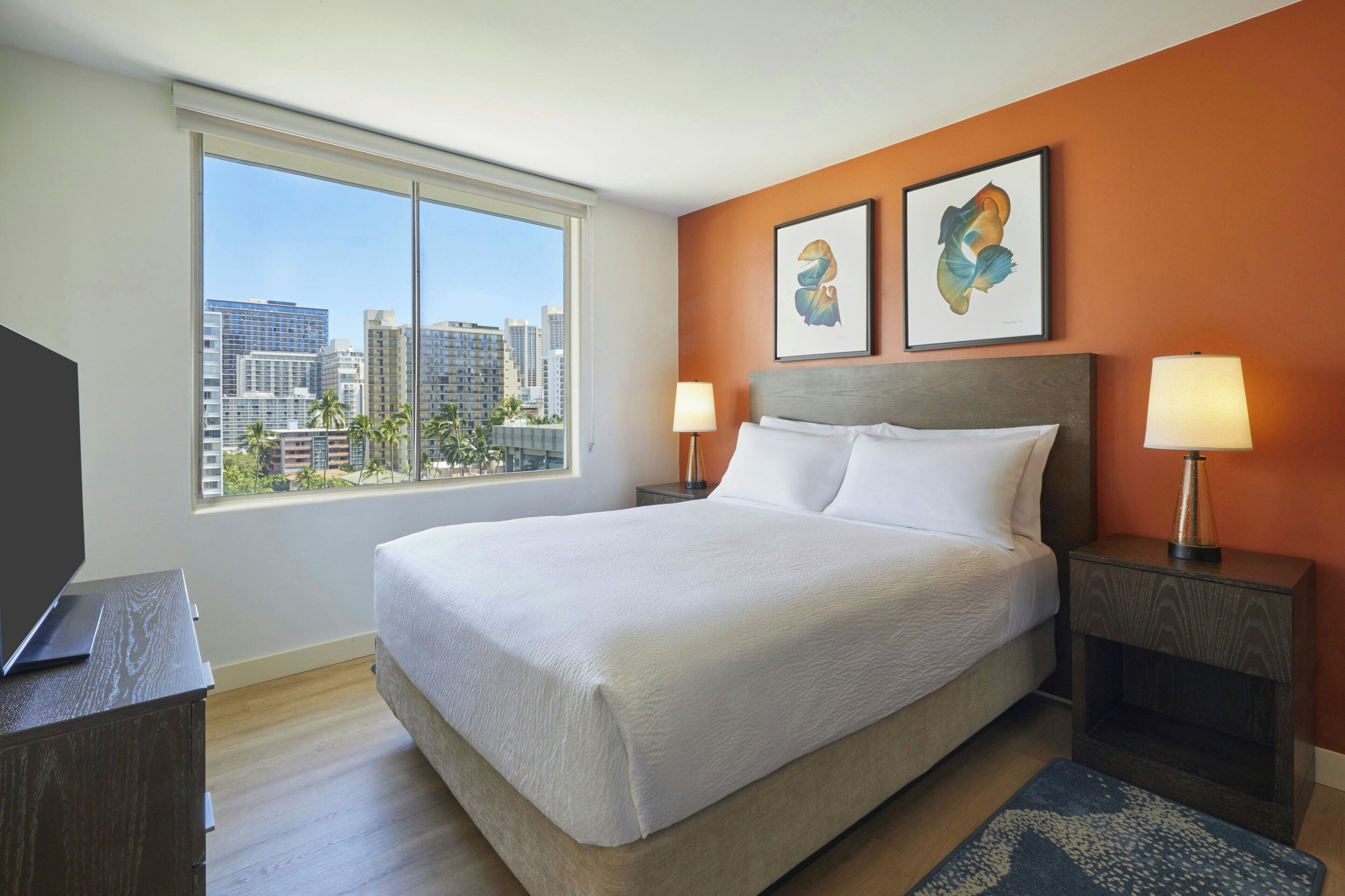 Habitación de hotel con una cama grande, dos mesitas de noche con lámparas, un televisor de pantalla plana sobre un aparador de madera y vistas a la ciudad a través de grandes ventanales.